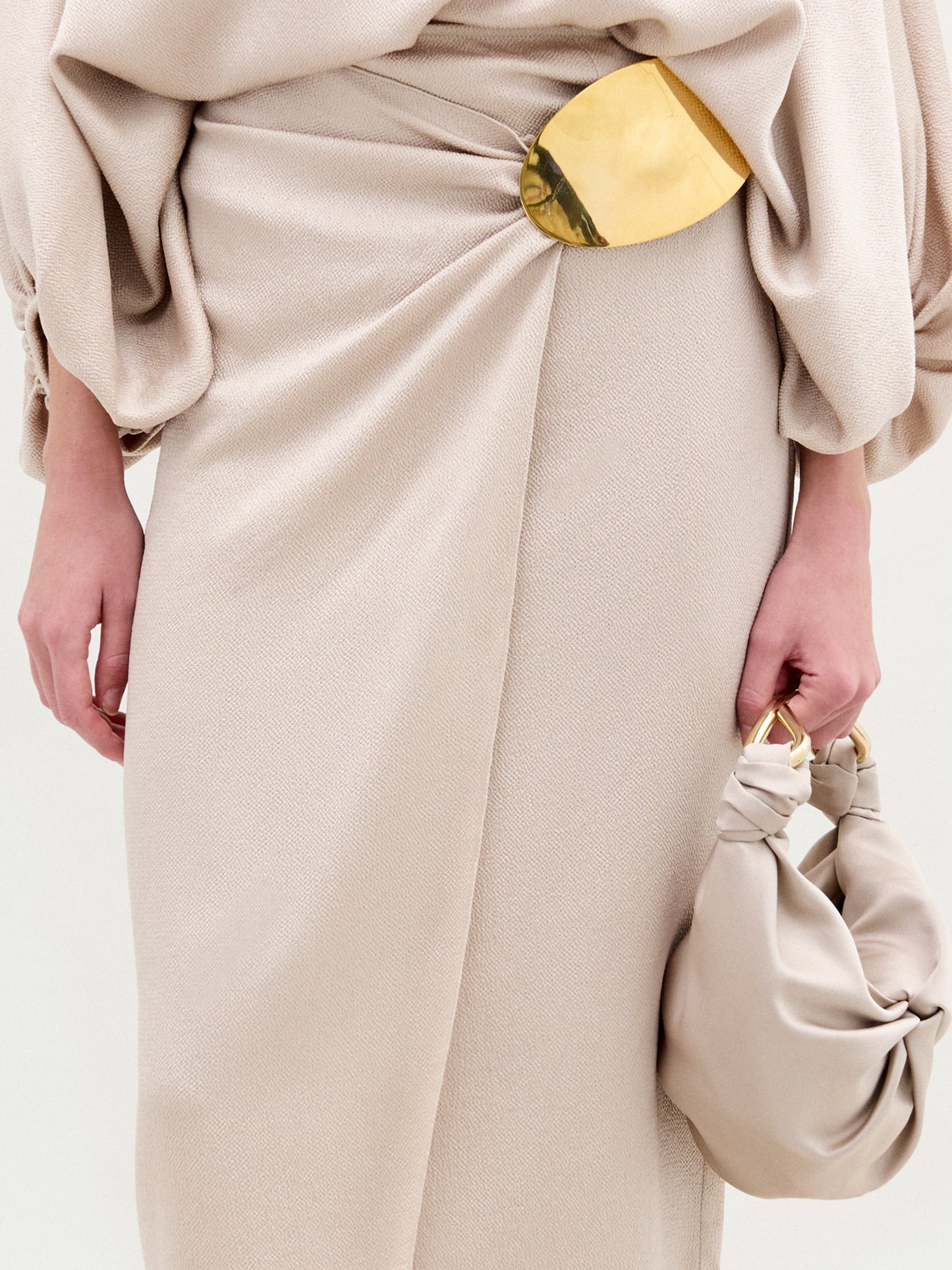 A person wears SVT DESIGNS' hammered satin beige Adrianne Skirt with a gold clasp at the waist, paired with a small beige handbag featuring knotted straps and gold-tone embellishments, showcasing its textured elegance.