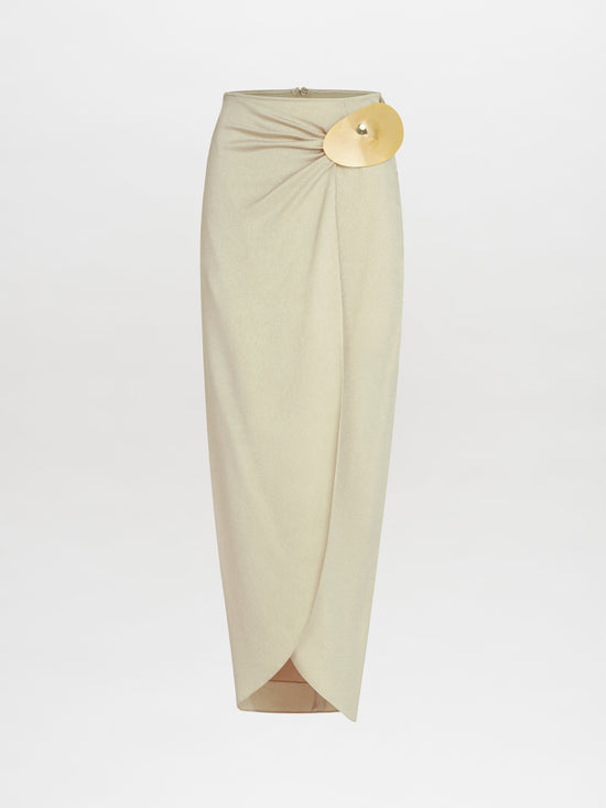 The Adrianne Skirt by SVT DESIGNS is a beige high-waisted midi crafted from hammered satin, featuring a draped design with a gold-tone embellishment at the waistband. Its elegant front overlap and long length create a sophisticated look against a plain background.