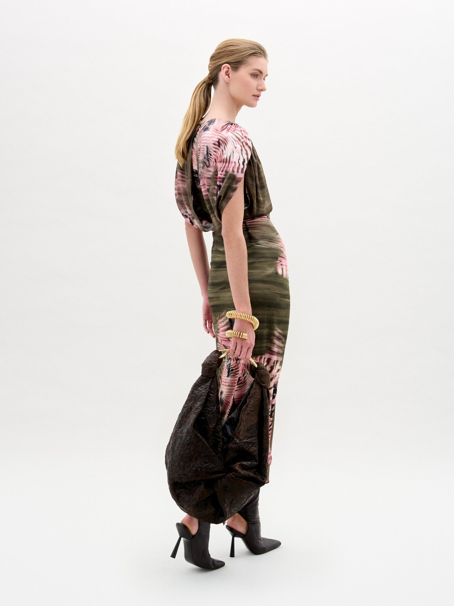 A person with long hair in a ponytail wears the Adrika Skirt by SVT DESIGNS, crafted from luxurious Italian fabric with green and pink patterns. They carry a large textured black bag and wear black high-heeled boots, all against a plain light gray background.