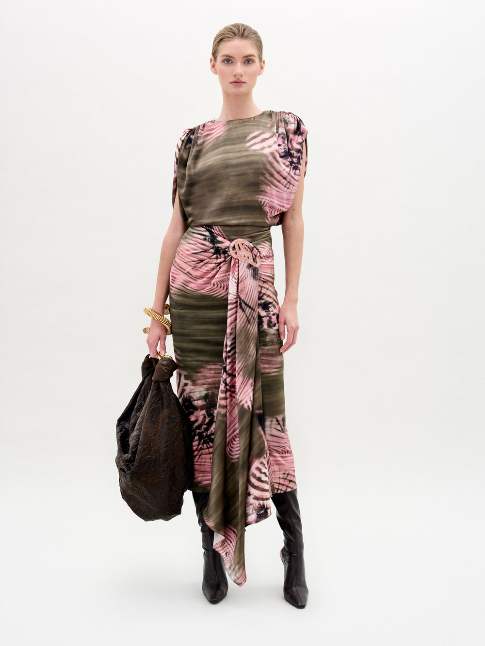 A woman stands against a plain background wearing the Adrika Skirt by SVT DESIGNS, featuring Italian fabric with pink, black, and olive green patterns. She pairs it with black boots, gold bracelets, and holds a large dark textured bag in her left hand.