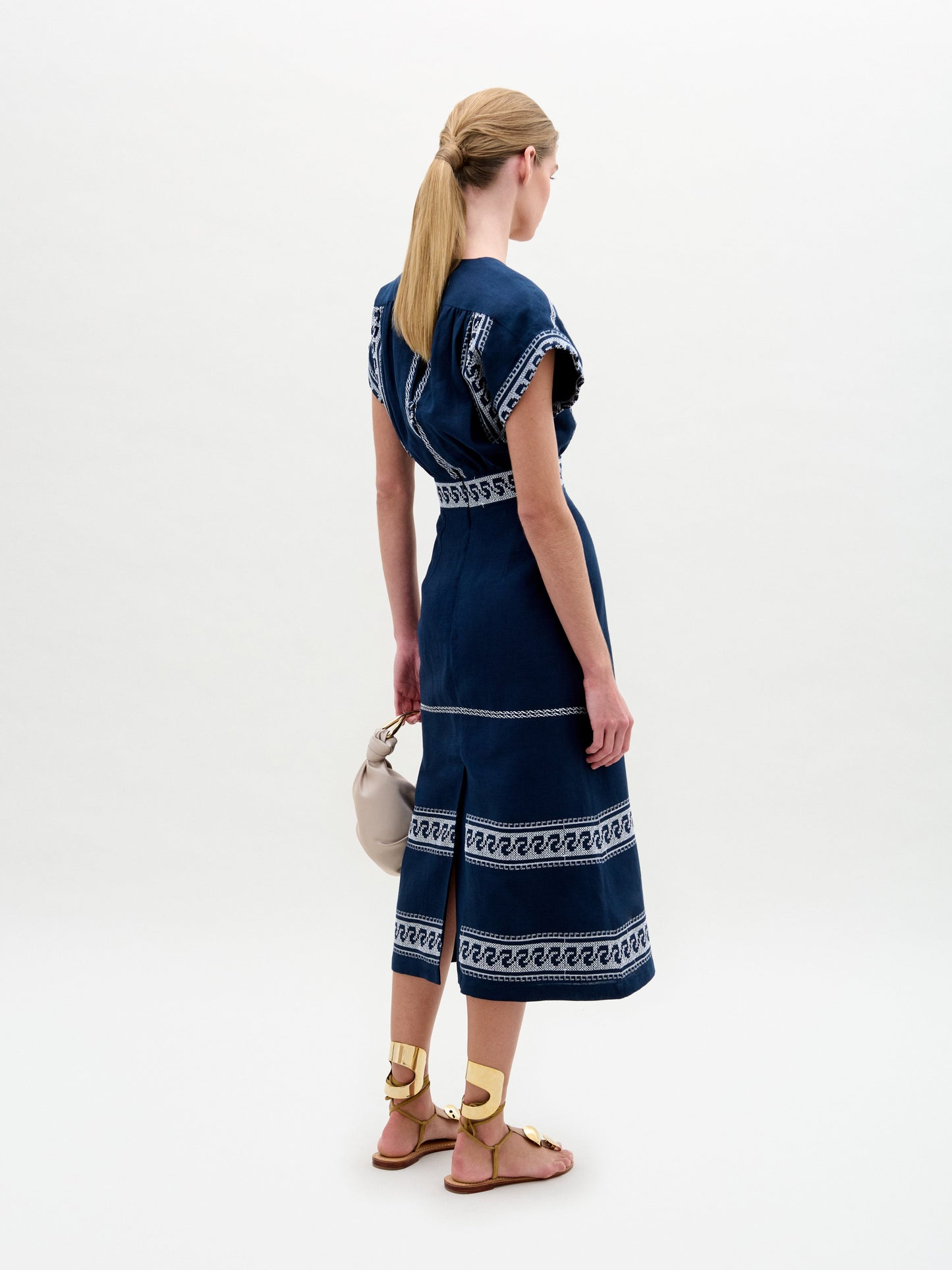 A person faces away, wearing the Ady Dress by SVT DESIGNS, a blue embroidered linen dress with white patterns. Their hair is in a ponytail, and they hold a beige handbag while wearing gold sandals. The backdrop is plain white.