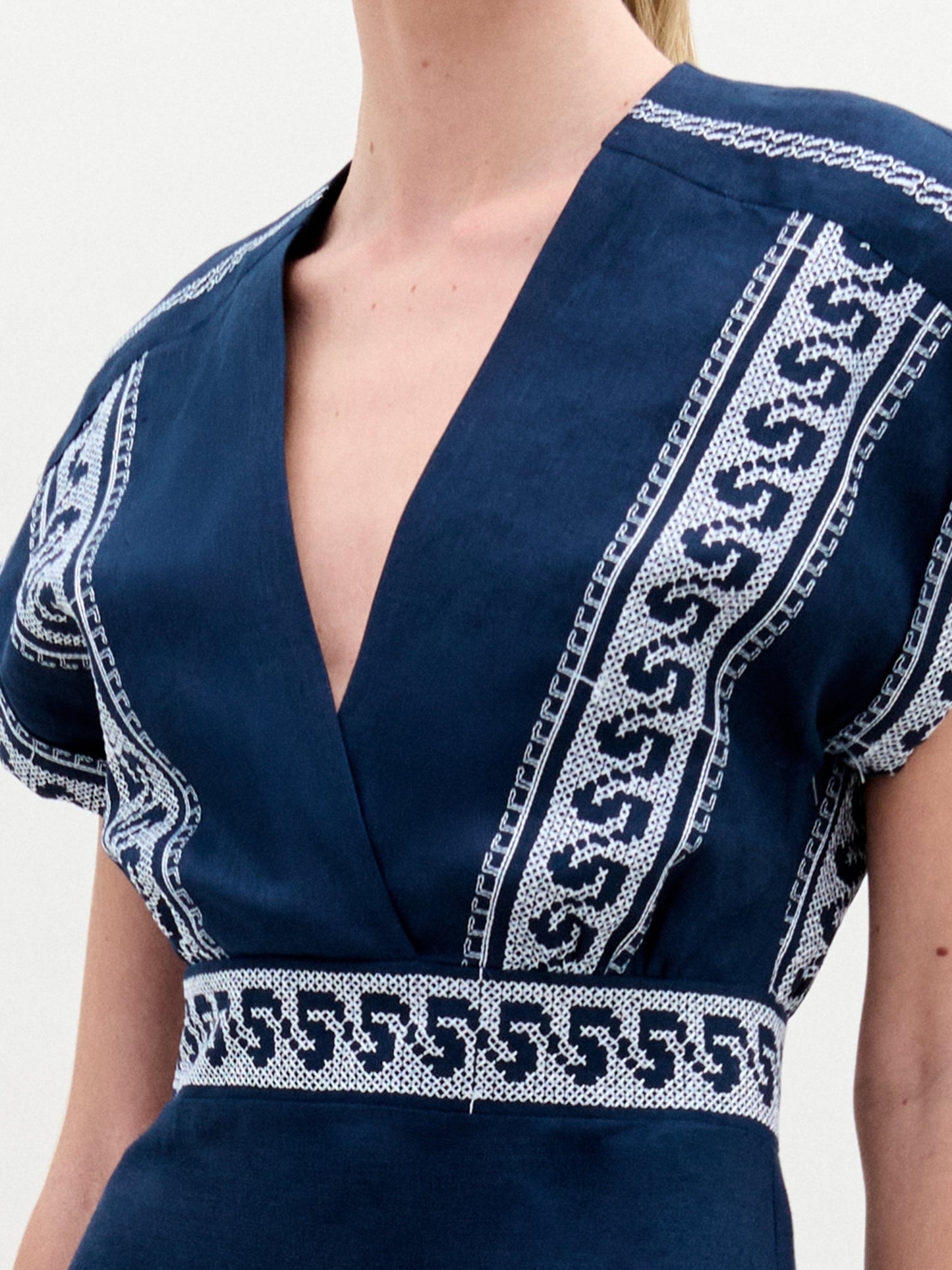 A person wears the SVT DESIGNS Ady Dress, a dark blue midi featuring a deep V-neckline with intricate white patterns and geometric designs on the shoulders and waist. Crafted from Italian fabric, the background is plain and neutral while their face remains unseen.