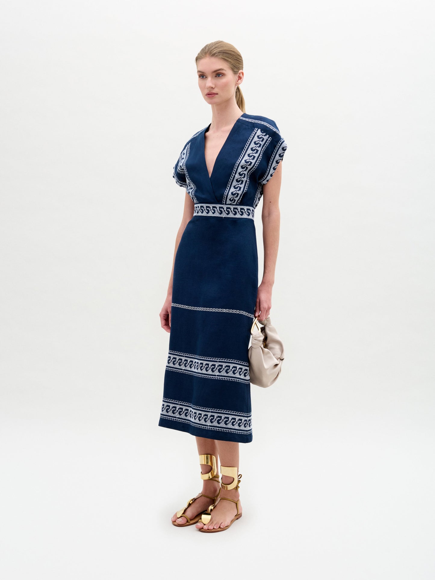 A woman poses against a plain backdrop wearing the SVT DESIGNS Ady Dress, a navy blue midi dress featuring white geometric patterns made from Italian fabric. She pairs it with gold sandals and carries a beige handbag. Her long hair is pulled back as she gazes forward neutrally.