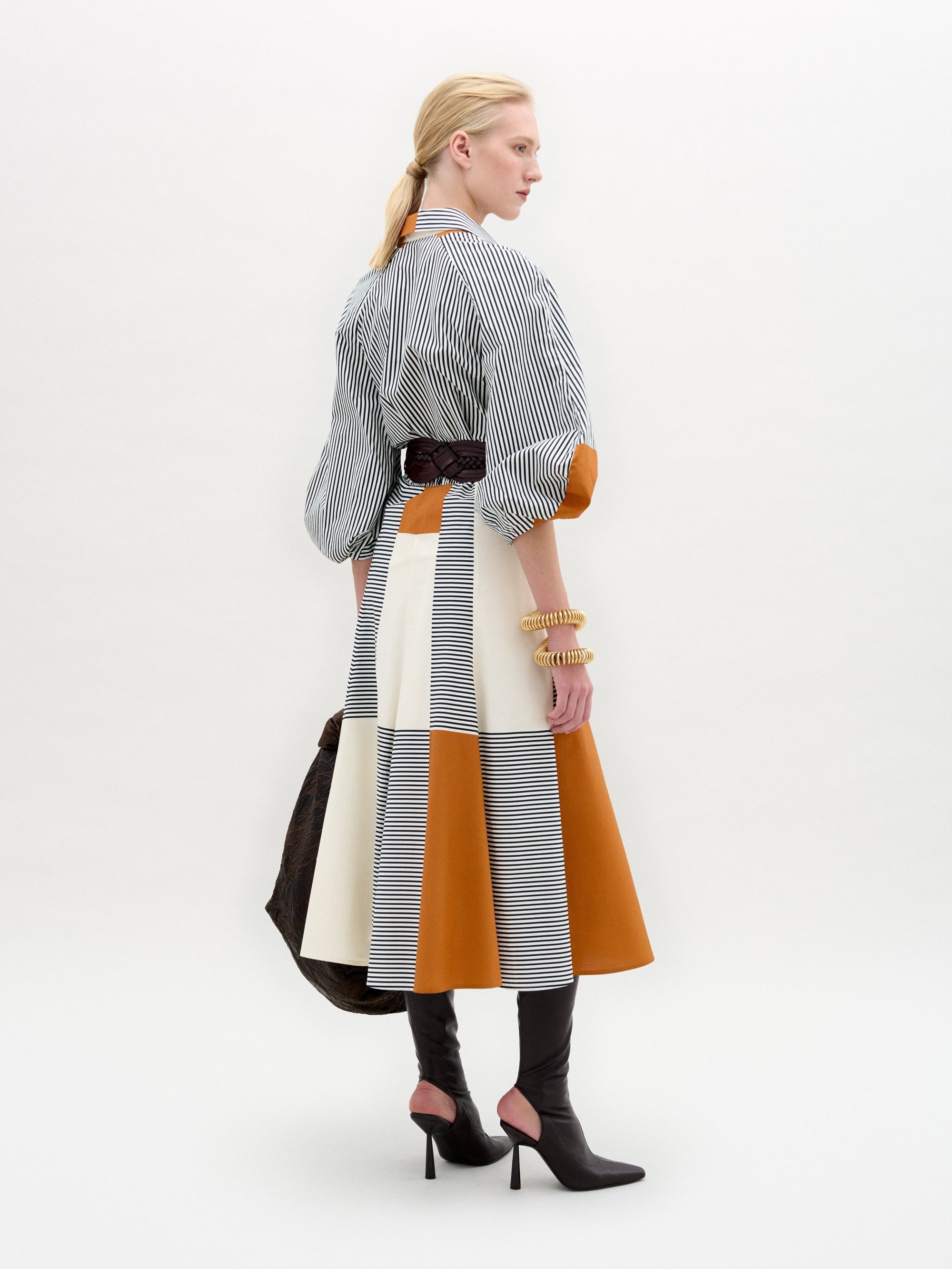 A woman stands against a white background wearing the Agostina Dress—striped and color-blocked, crafted from Italian fabric—paired with high brown boots, a wide belt, and holding a large dark bag.