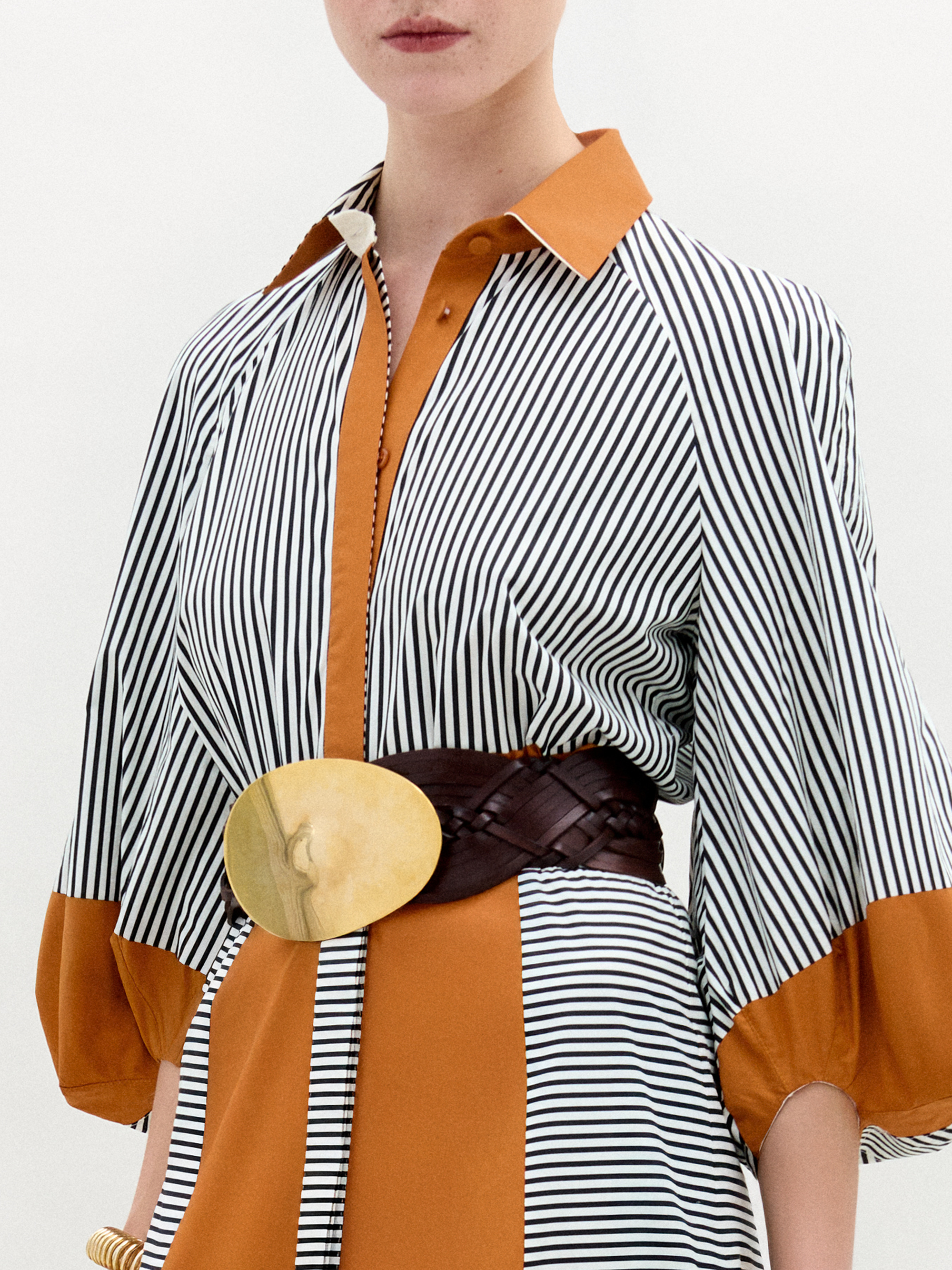 The Agostina Dress by SVT DESIGNS, made from fine Italian fabric, showcases rust accents and a gold circular belt buckle. It features wide sleeves and a pointed collar. Only the person's lips and neck are visible in the frame while wearing this striped midi dress.