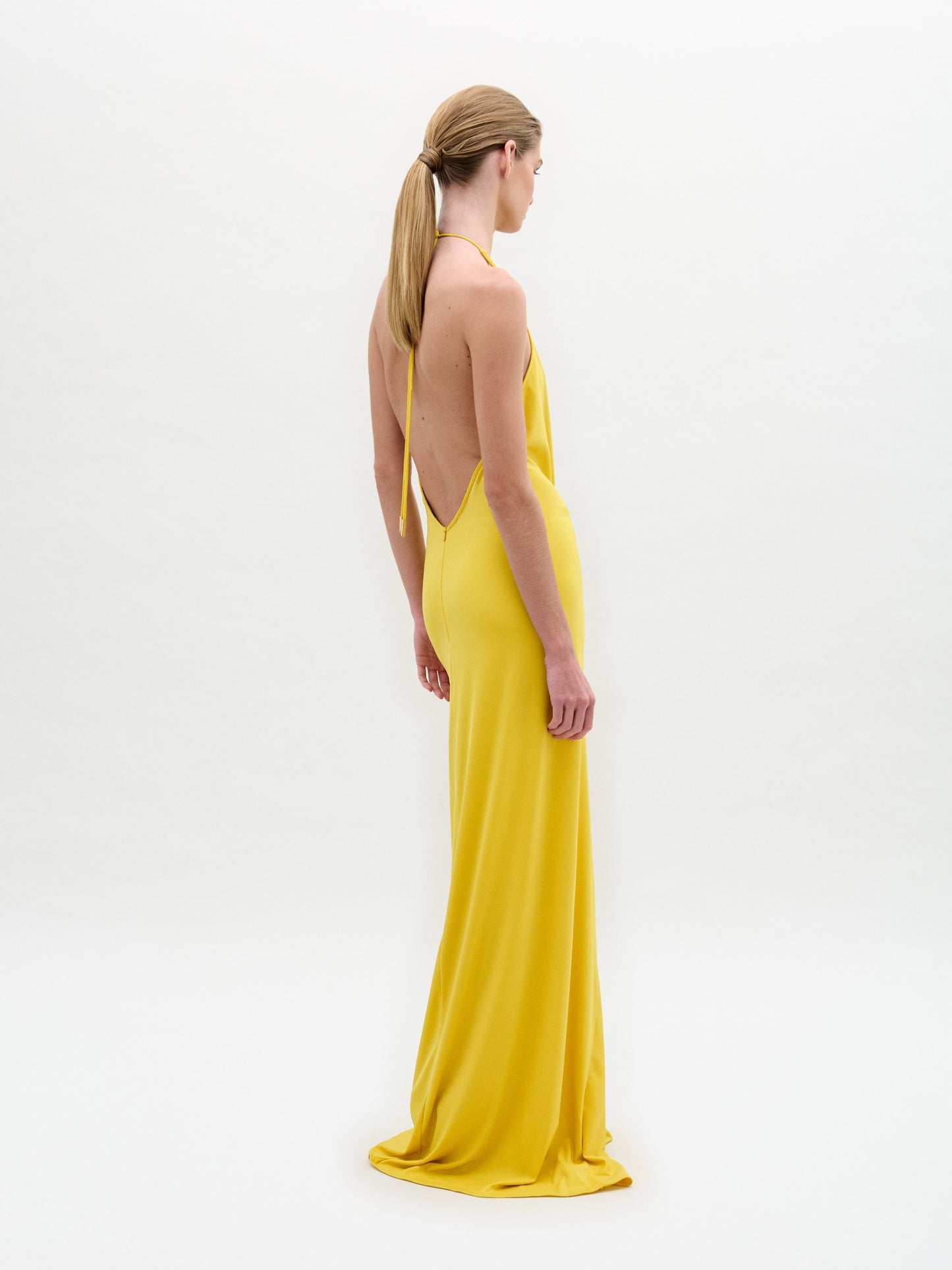 A person with long hair tied back models an elegant SVT DESIGNS Aiko Dress, a yellow backless halter maxi gown made from luxurious Italian fabric. The floor-length dress is set against a plain off-white background as the model turns slightly to the side, facing away from the camera.