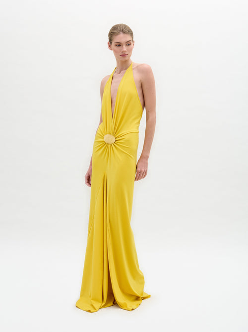 A person stands wearing the Aiko Dress by SVT DESIGNS; it's a yellow gown made from luxurious Italian fabric, featuring a halter neckline with central embellishment. The stunning maxi dress has a floor-length skirt with an elegant slit, set against a plain white background.