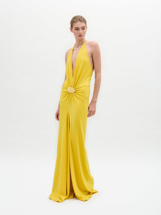 A person stands wearing the Aiko Dress by SVT DESIGNS; it's a yellow gown made from luxurious Italian fabric, featuring a halter neckline with central embellishment. The stunning maxi dress has a floor-length skirt with an elegant slit, set against a plain white background.