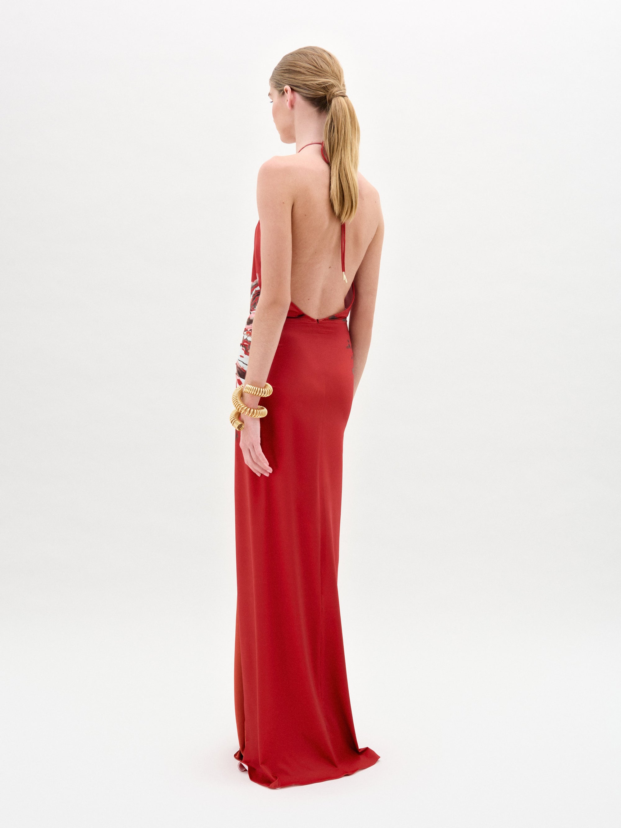 A person with long hair stands against a plain white background, wearing the Aiko Dress by SVT DESIGNS. This elegant red maxi dress crafted from luxurious Italian fabric features a backless design, gold embellishments, a halter neck, and intricate side detailing.