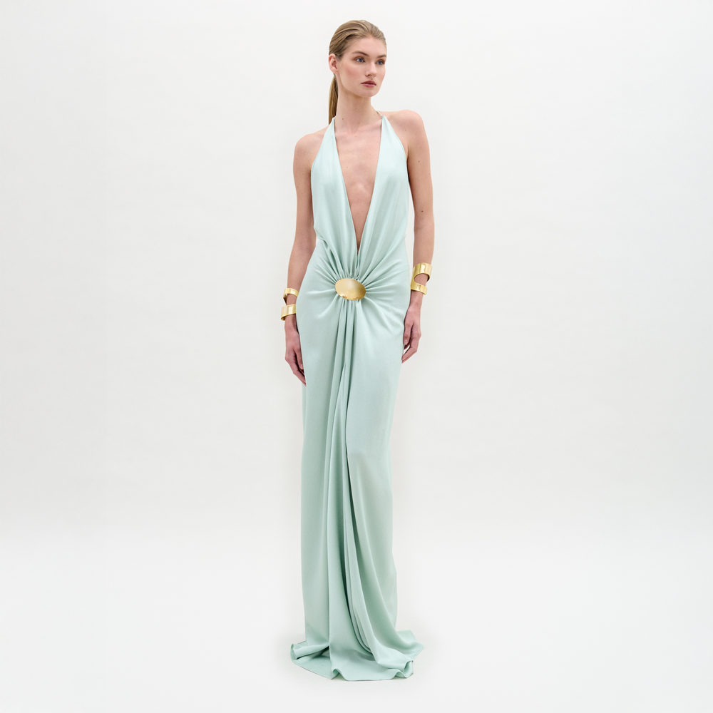 A woman stands against a white background wearing the Aiko Dress by SVT DESIGNS, a light blue maxi with a deep plunging neckline and gathered waist, crafted from luxurious Italian fabric and paired with gold cuff bracelets.