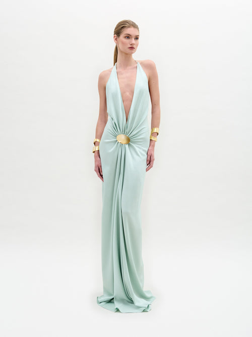 A model poses in the SVT DESIGNS Aiko Dress, a light green, floor-length gown made from exquisite Italian fabric. It features a plunging neckline and central gathered design, accessorized with gold embellishments on her wrists and her hair elegantly pulled back against a plain backdrop.