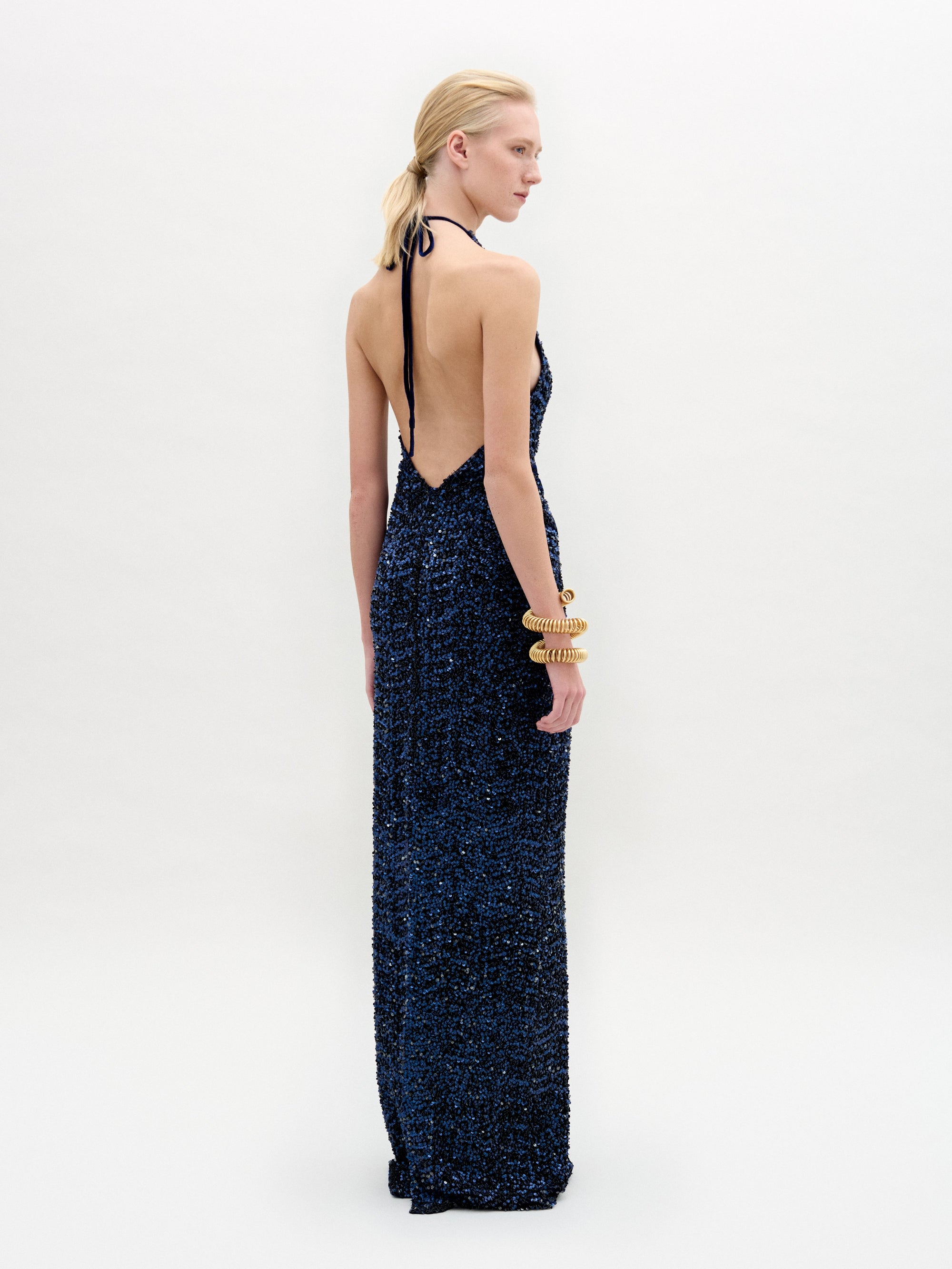A profile shows someone in the elegant Aiko Dress by SVT DESIGNS, crafted from luxurious Italian fabric. The dark, textured halter-neck gown features an open back. Their hair is tied back, complemented by large chunky bracelets on one wrist against a plain white background.