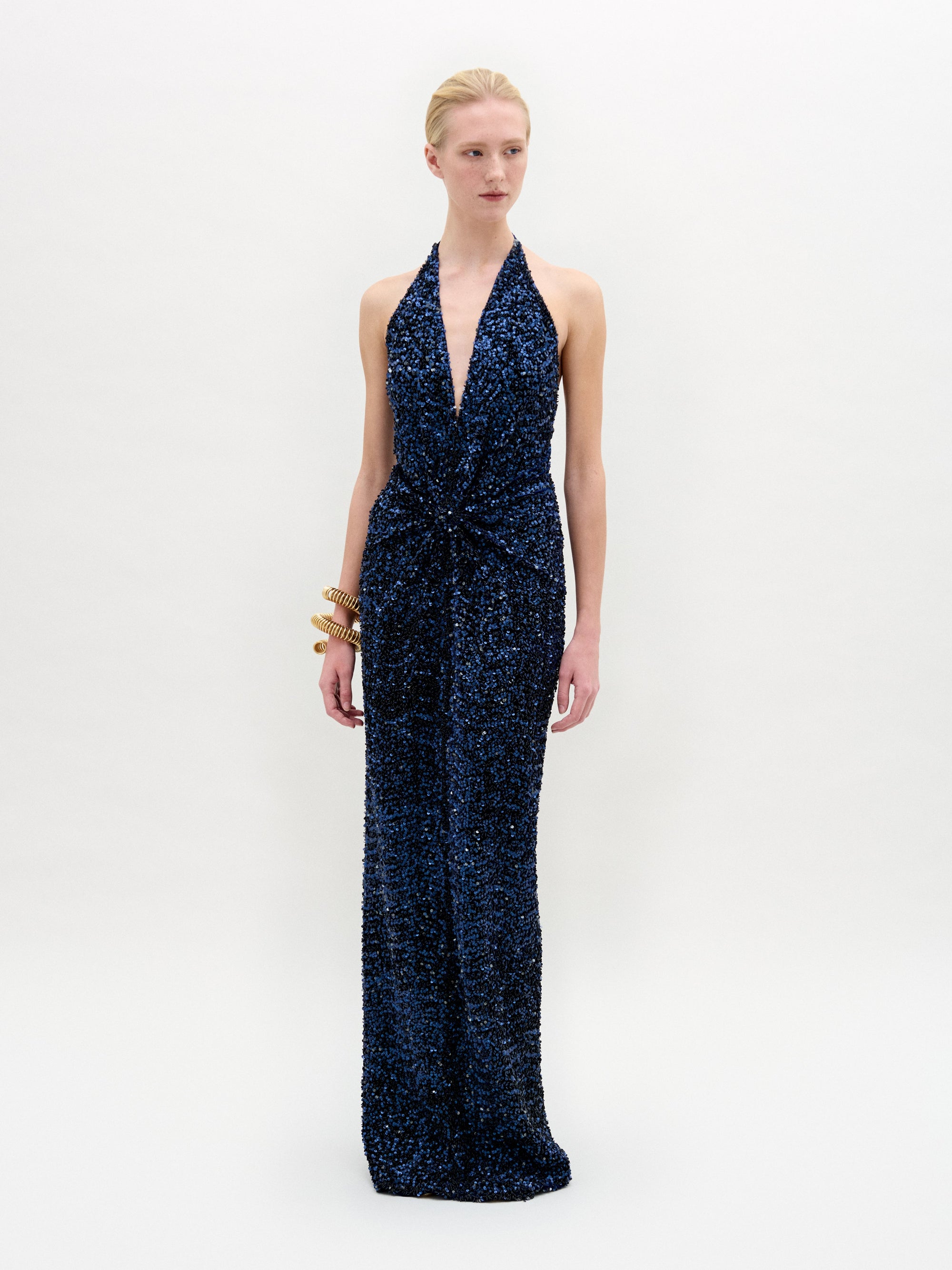 A person wears the sleek, sleeveless Aiko Dress by SVT DESIGNS in dark blue sequined Italian fabric with a deep V-neck, set against a plain white background. They accessorize with chunky gold bangles on the left wrist and have their hair styled neatly back.
