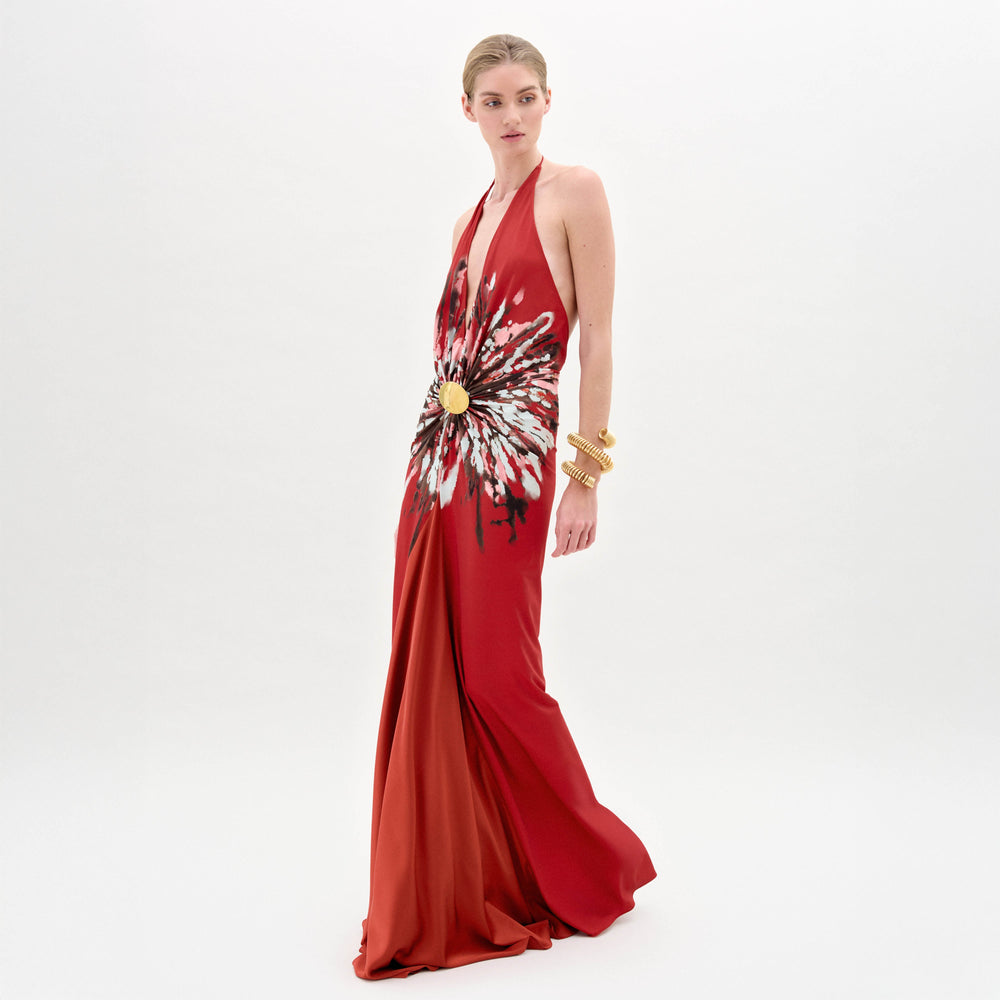 A woman stands against a white background wearing the Aiko Dress by SVT DESIGNS—a red halter-neck maxi with an abstract floral waist detail—paired with gold bracelets. Her hair is slicked back as she looks slightly left.