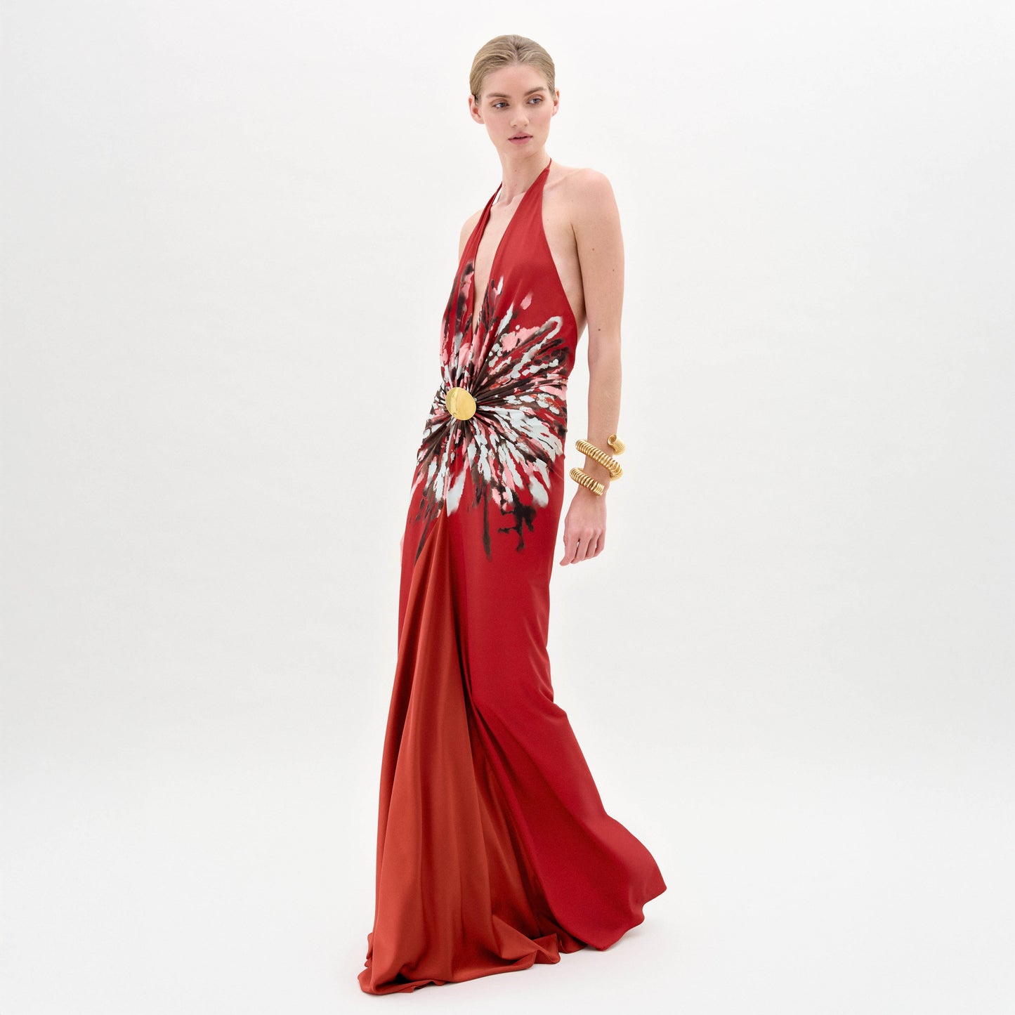 A woman wearing the Aiko Dress—a sleeveless red maxi with a bold white and black burst pattern at the waist and gold embellishments—stands against a plain white background, accessorized with multiple gold bracelets.