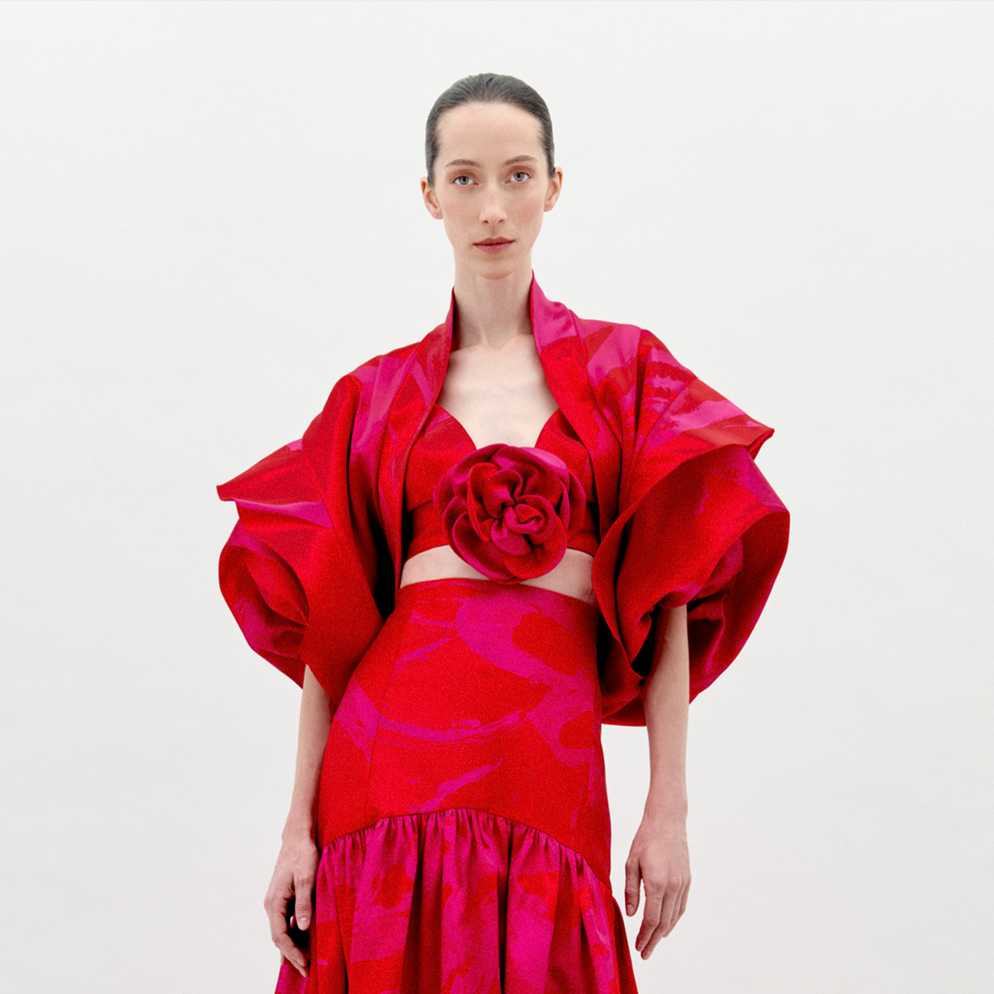 A woman stands against a plain white background wearing the SVT DESIGNS Aleen Shrug—an oversized red and pink patterned piece with voluminous sleeves and a large flower detail at the center—facing forward with a neutral expression.