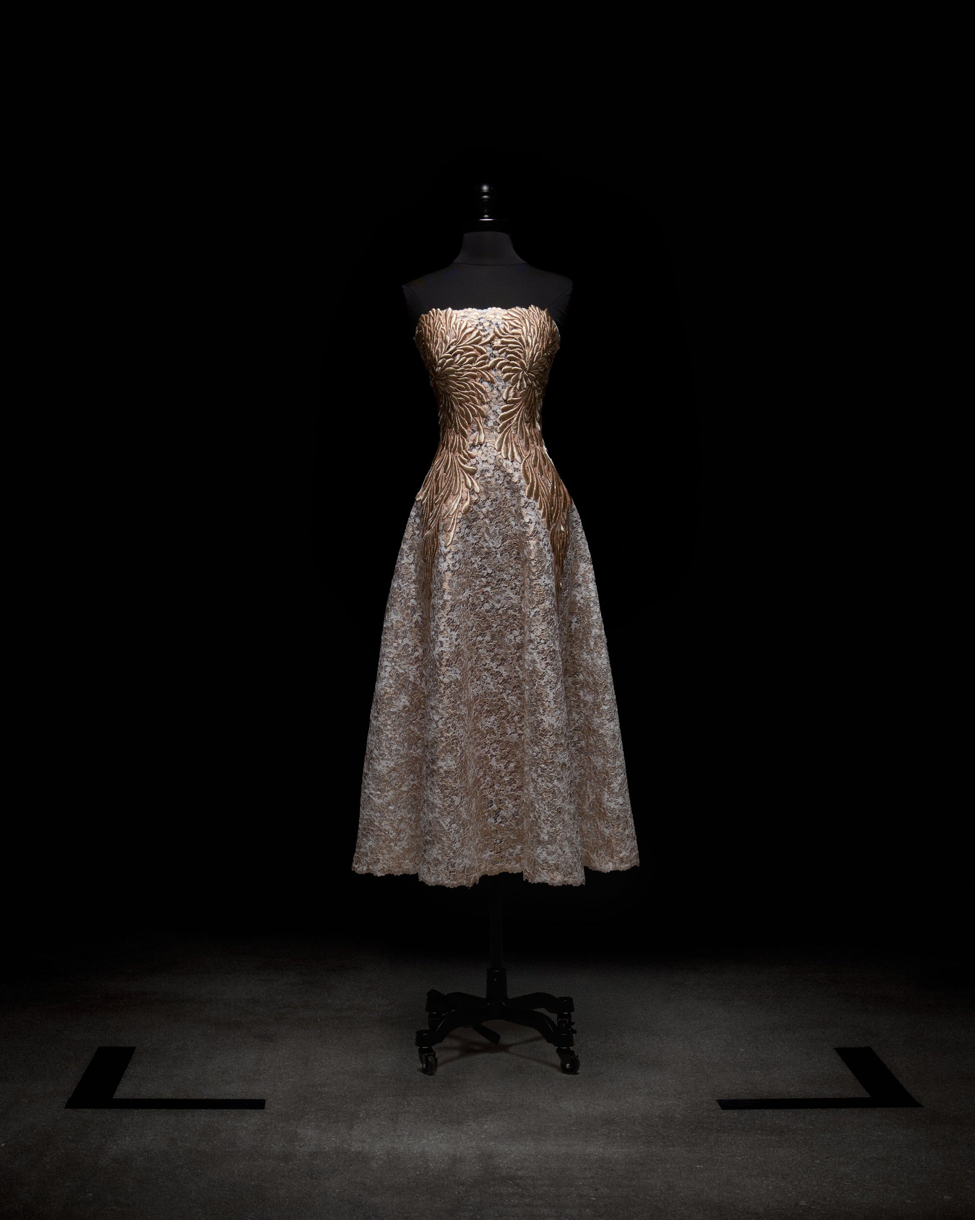 The Alejandra Dress by SVT DESIGNS, showcased on a black mannequin against a dark backdrop, has a strapless fitted gold bodice with a swirling pattern and is complemented by a full light gray lace skirt that falls to mid-calf.