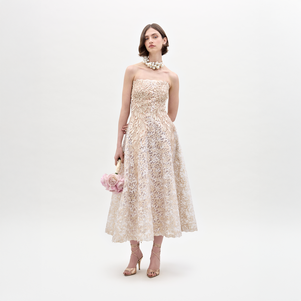 A woman models the Alejandra Dress by SVT DESIGNS, a cream-colored strapless guipure lace midi, against a white background. She accessorizes with strappy heels, a chunky pearl necklace, and a small pink flower-shaped purse.