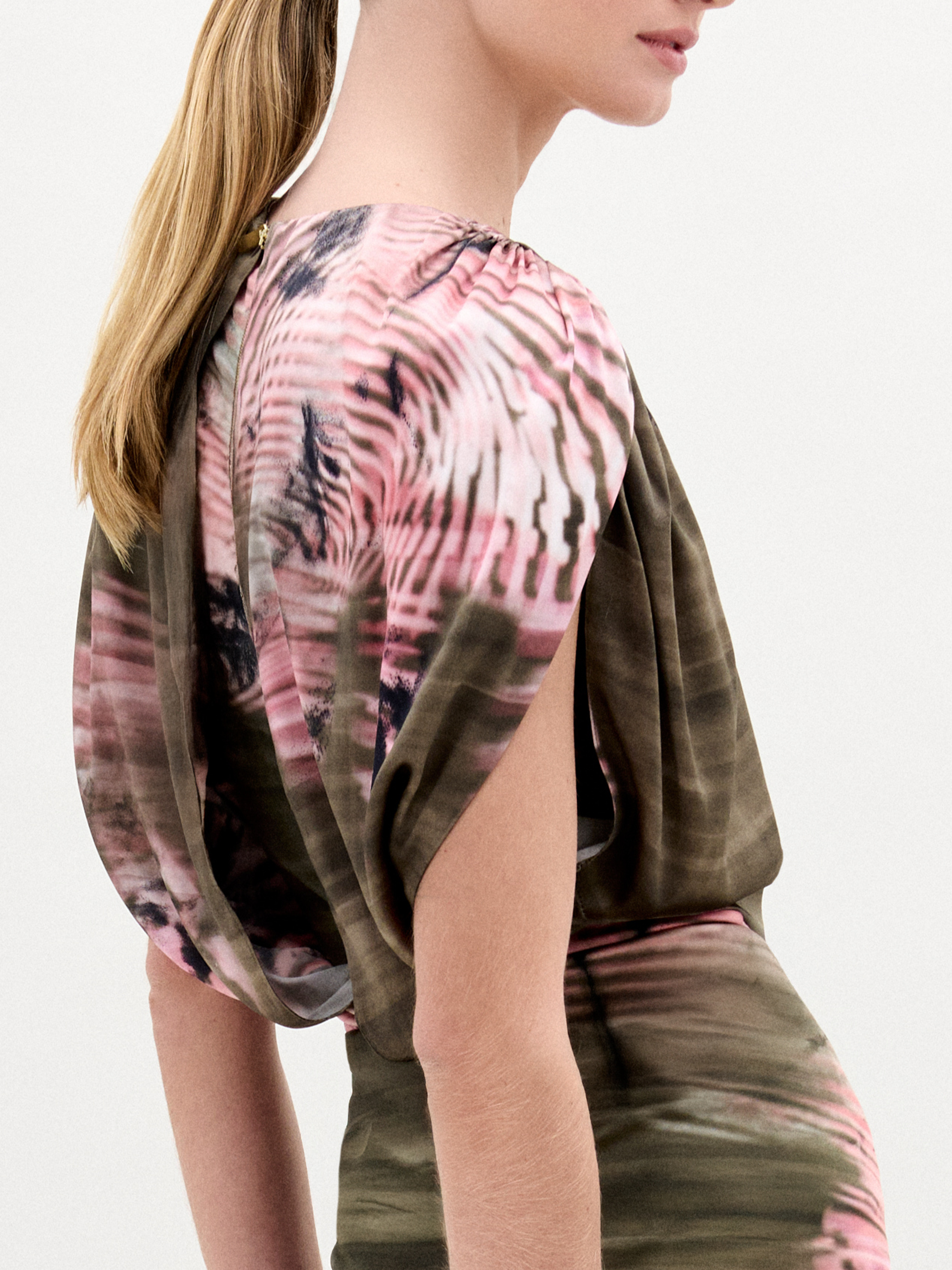 A woman stands against a plain background wearing the Alisa Blouse, featuring a pink and olive green pattern crafted from Italian fabric. She holds a brown bag and wears gold bracelets.