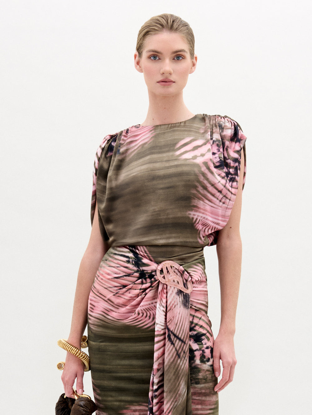 A woman stands against a plain light background wearing the Alisa Blouse by SVT DESIGNS, featuring Italian fabric with a pink and brown abstract pattern. She holds a brown bag, wears chunky gold bracelets on her left wrist, and has her hair pulled back.