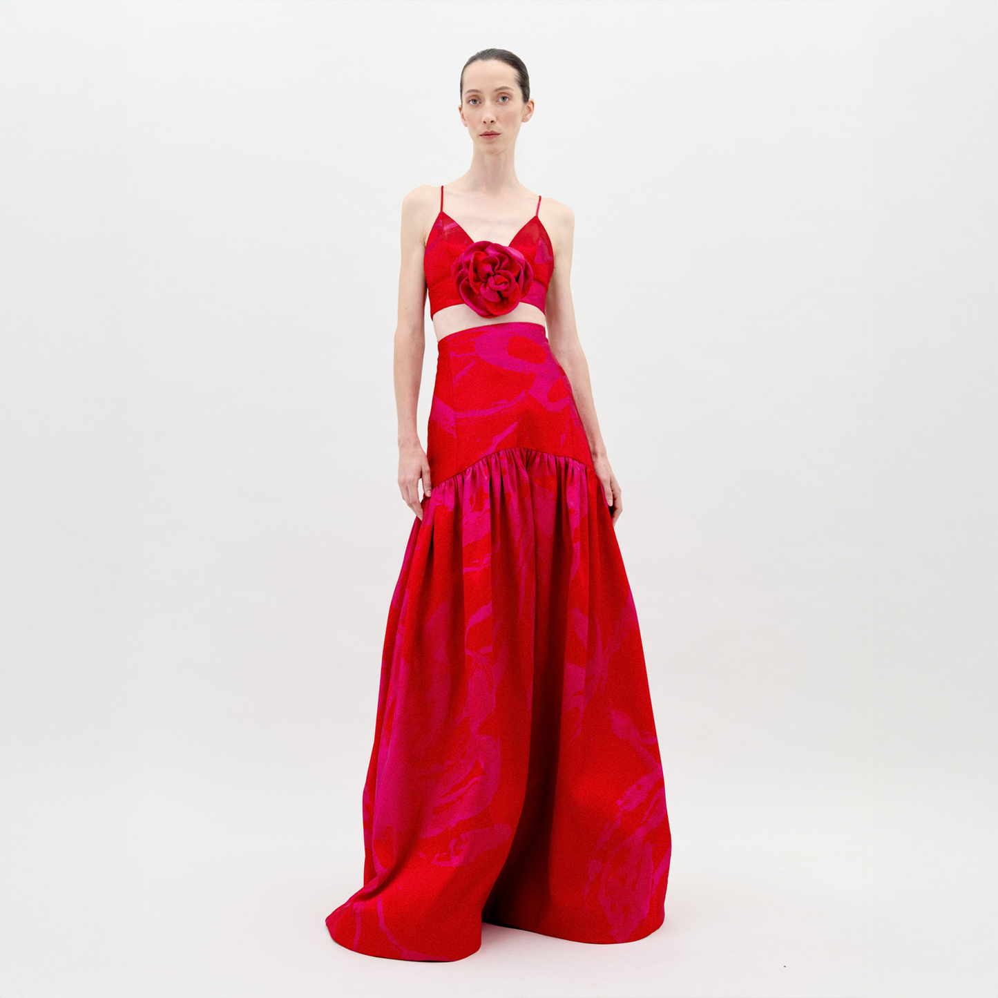 A person poses against a plain white background in the SVT DESIGNS Helen Dress Set, featuring a vibrant red floor-length taffeta skirt, fitted bodice with thin straps, and a bold floral detail at the chest for a voluminous silhouette.