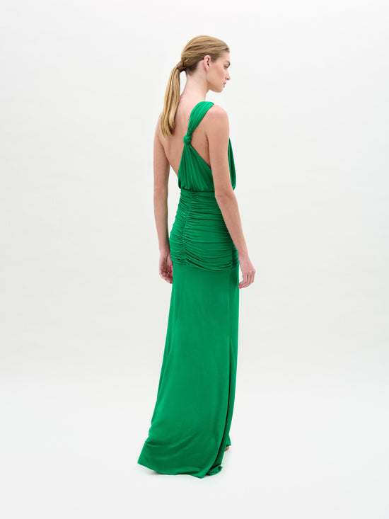 A woman on a white background wears the Antara Dress by SVT DESIGNS, a green maxi crafted from soft jersey. It has a draped design with back ruching and an asymmetric neckline. Her hair is in a low ponytail as she looks left.