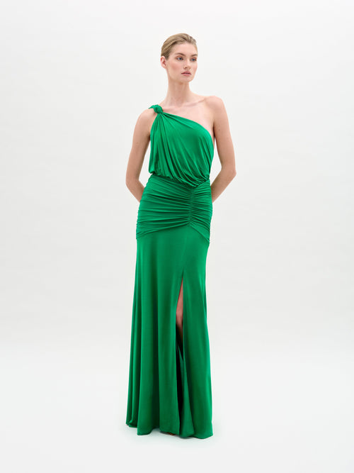 The individual stands against a white background, wearing the Antara Dress by SVT DESIGNS—a green jersey maxi dress with an asymmetric neckline, gathered waist design, and side slit. Their hands are clasped in front of them.