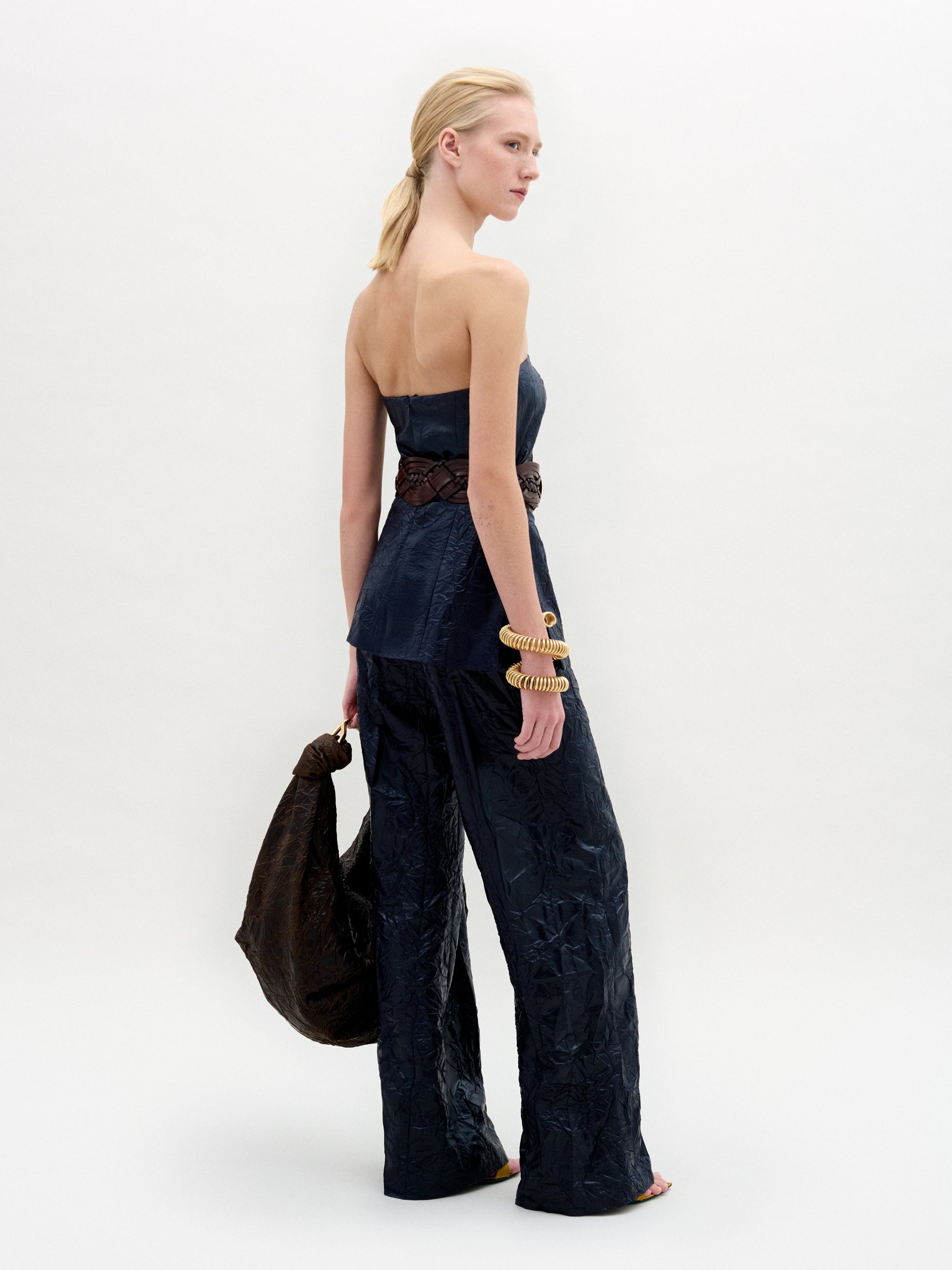 A model poses against a plain backdrop in SVT DESIGNS' Emine Pant, featuring matching high-waisted, wide-leg pants and a dark strapless top made from luxurious Italian fabric. She accessorizes with large bracelets, holds a textured bag, and wears her hair tied back simply.