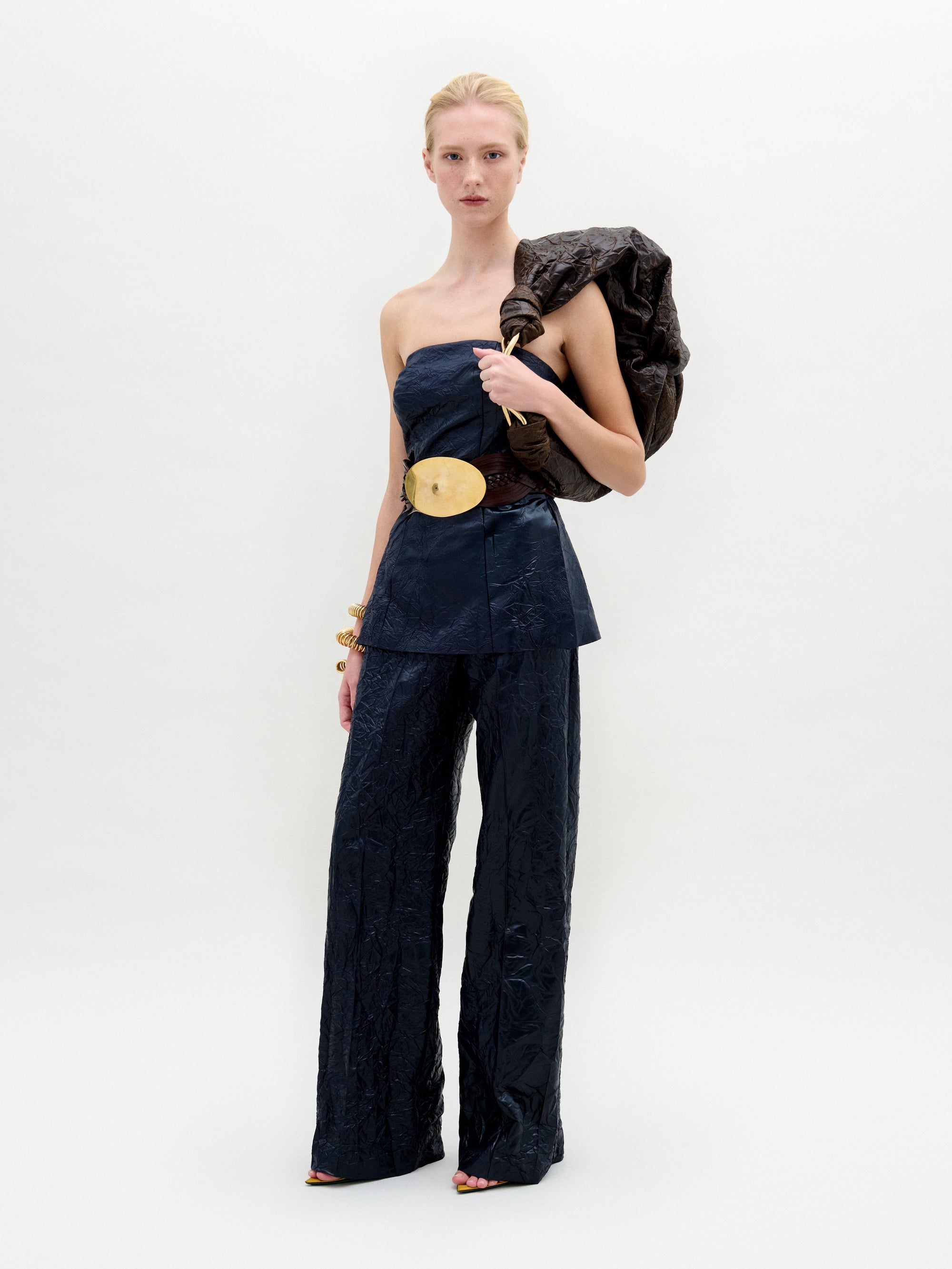 A person poses against a plain background in SVT DESIGNS' Emine Pant, featuring a navy strapless top and wide-leg silhouette crafted from luxurious Italian fabric, accessorized with a large gold belt. They hold a textured jacket over their shoulder and wear black open-toe heels.