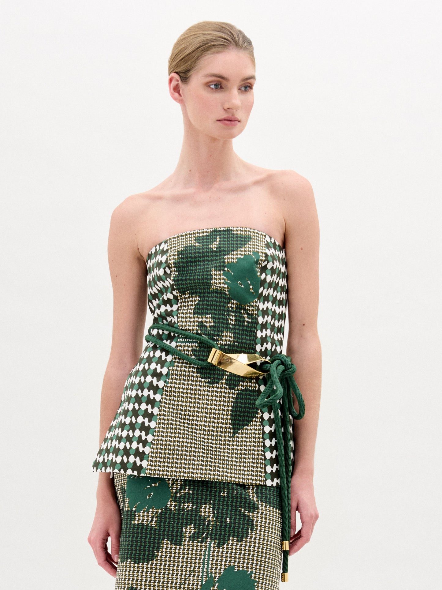 A woman with blonde hair in a low bun wears the SVT DESIGNS Astor Top, a strapless bustier in green, beige, and white Italian fabric, styled with a matching skirt and green belt with gold accents against a plain white background.