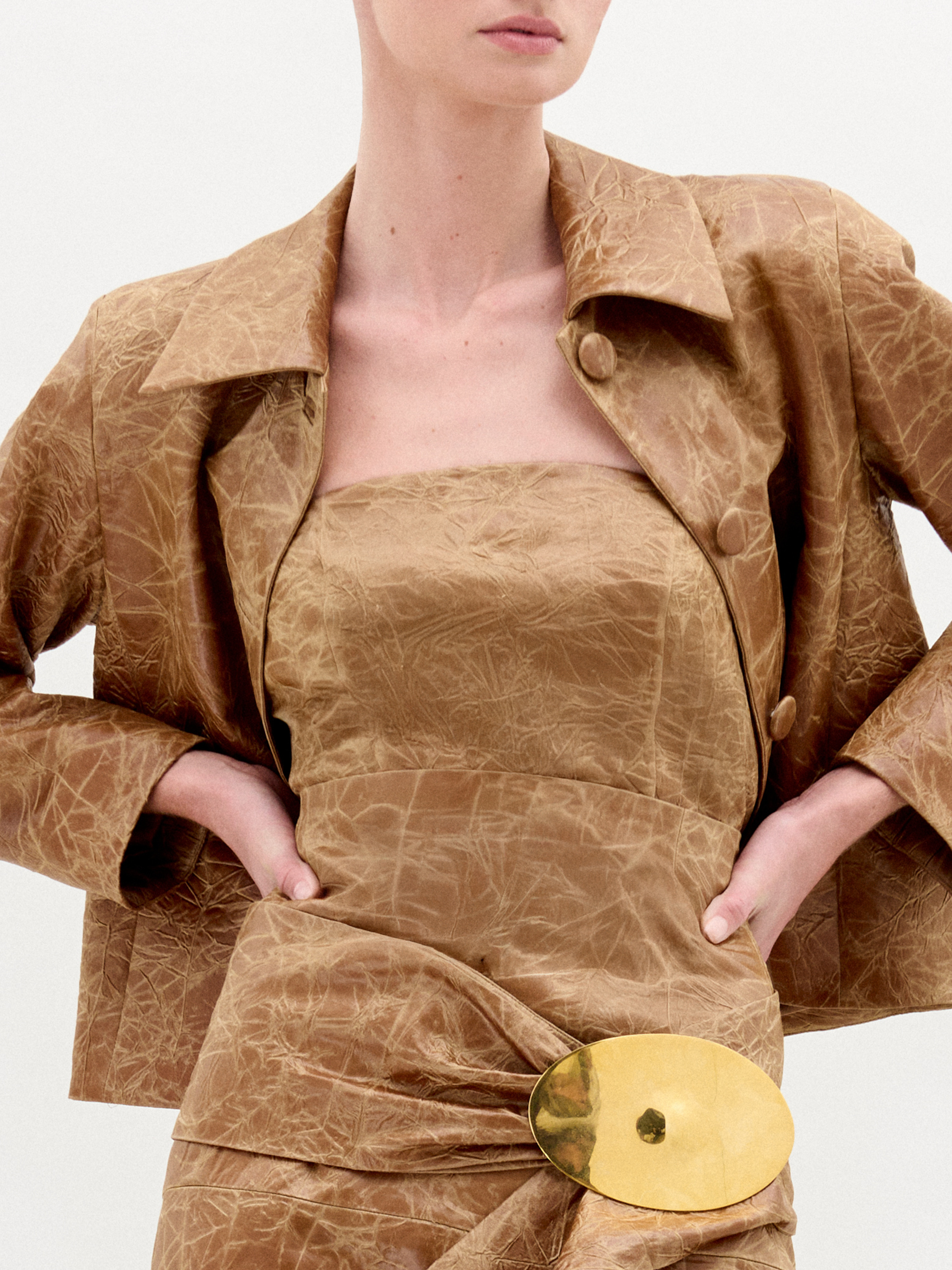A woman wearing the Astor Top, a crinkled brown strapless style with a fitted waist and large gold belt buckle, stands against a plain white background.