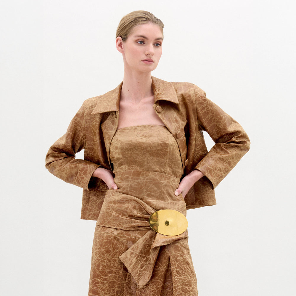 A woman wearing the Astor Top, a crinkled brown strapless style with a fitted waist and large gold belt buckle, stands against a plain white background.