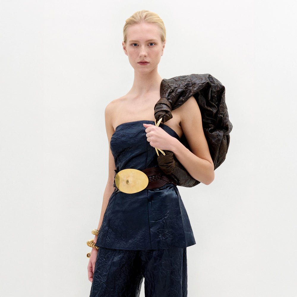 A woman in the navy Astor Top and matching pants poses with a large brown textured bag over her shoulder, styled with a fitted waist, wide belt featuring a large oval buckle, and gold bracelets.