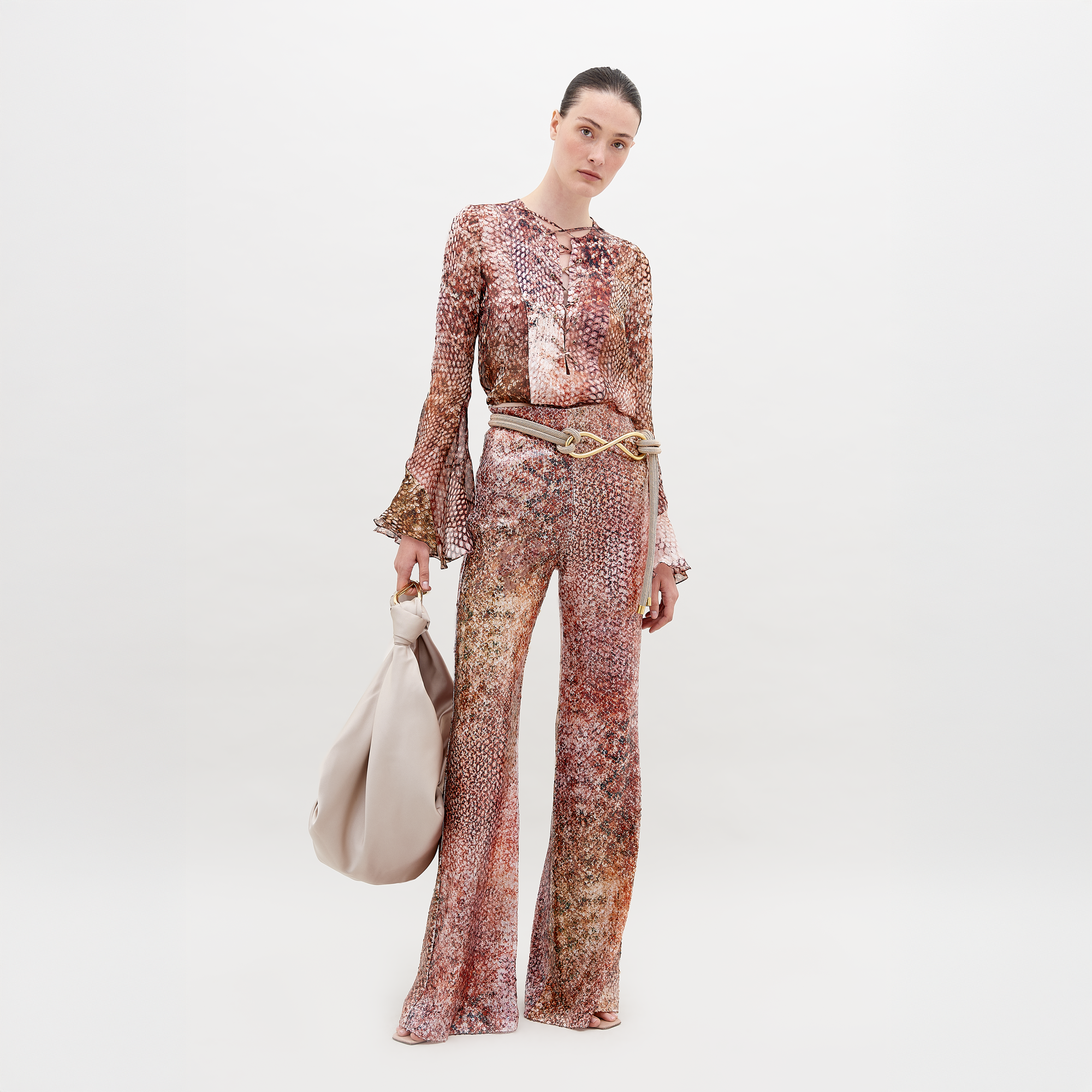 A person poses against a white background wearing high-waisted Avellino Pant Peach Animal Print by SVT DESIGNS, paired with a multicolored long-sleeved top. They hold a large, light-colored handbag, exuding Italian textile elegance.