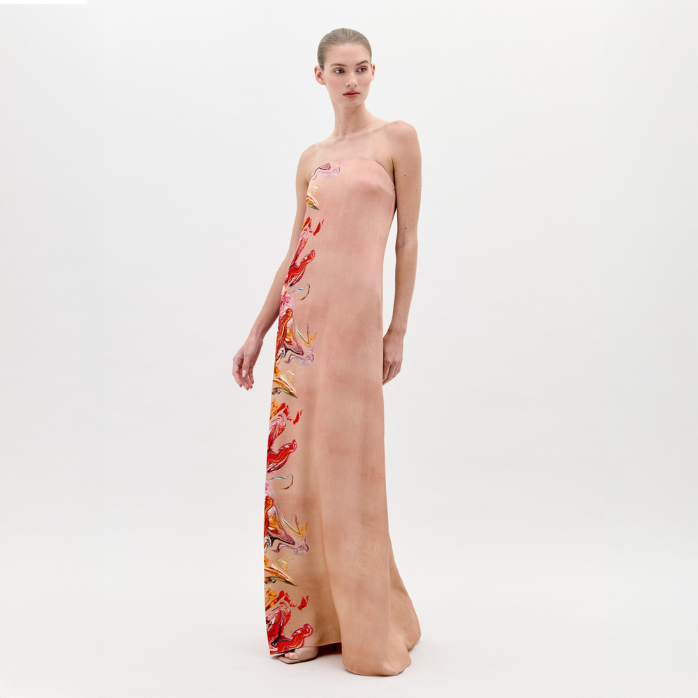 A woman models the Adalgisa Dress by SVT DESIGNS against a white background. The long, strapless maxi dress features red, yellow, and purple abstract designs along one side. She gazes slightly aside with one arm relaxed by her side.