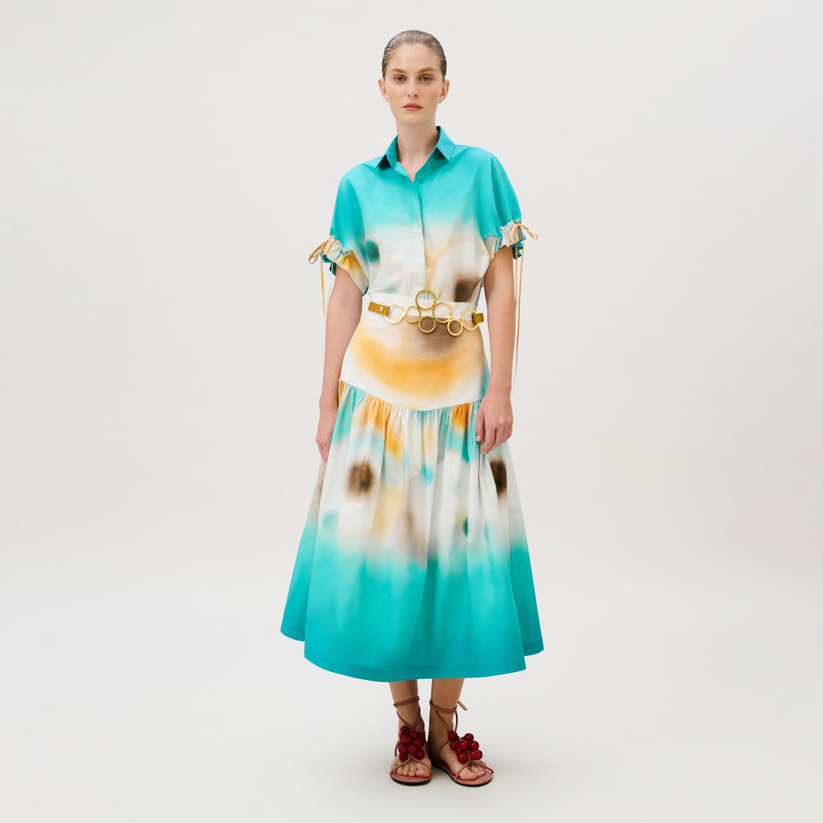 A person wearing an Aliana Skirt midi dress in a turquoise and yellow tie-dye cotton, featuring a gold-toned circular belt and lace-up sandals with red decorative accents.
