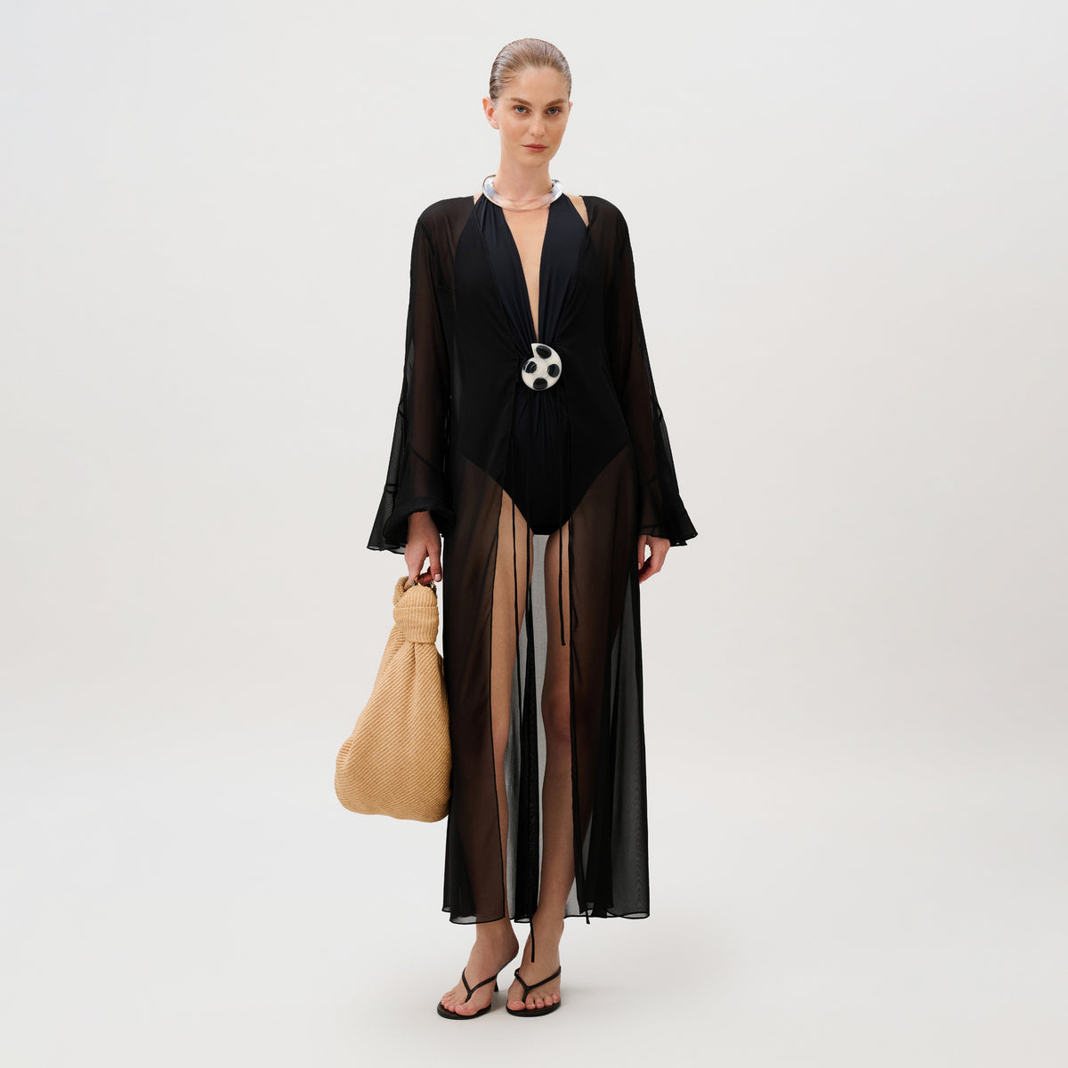 A woman wears a black swimsuit under an Amaris Tunic made of sheer chiffon with a central circular clasp, carrying a large tan woven handbag.
