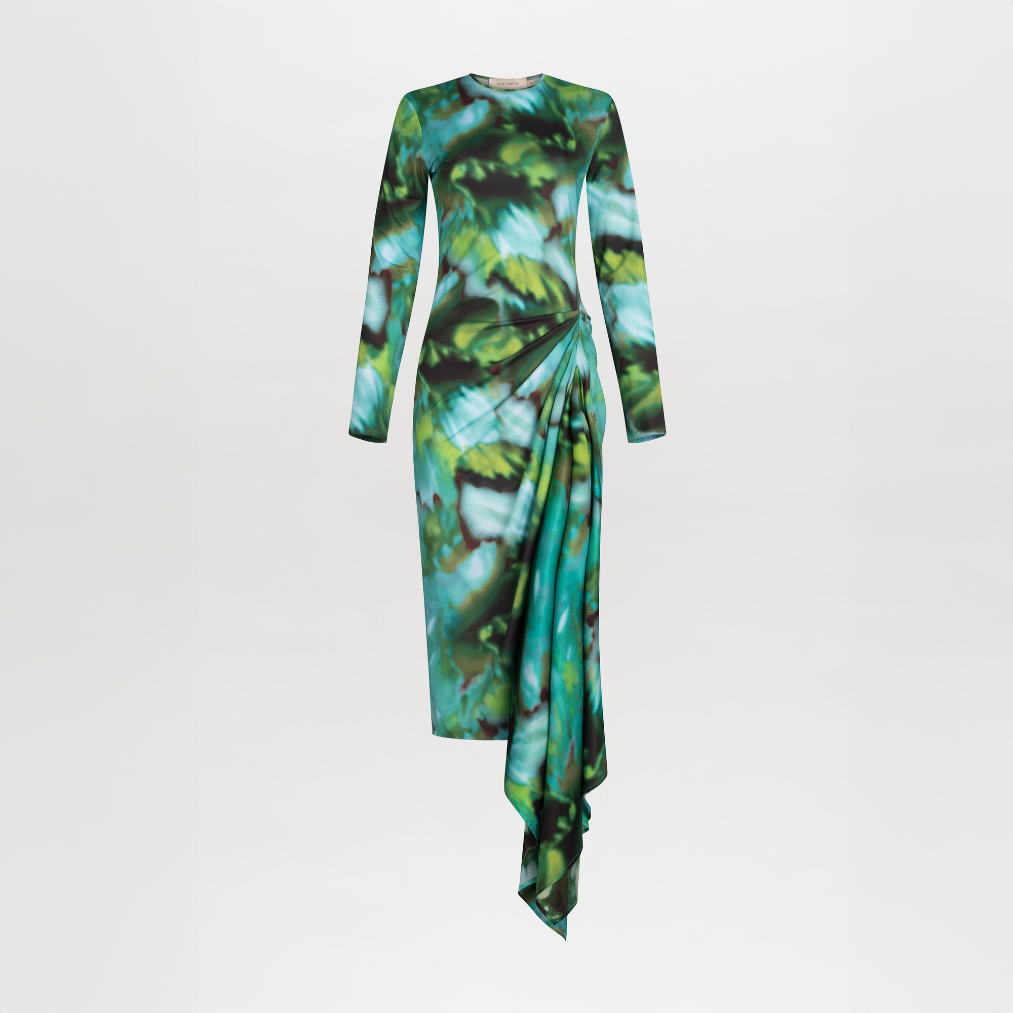 The Ananya Dress by SVT DESIGNS is a long-sleeved midi dress in green and blue leaf prints, made from 100% viscose. It features a gathered drape detail, an asymmetrical skirt, and a striking hem on a white background.