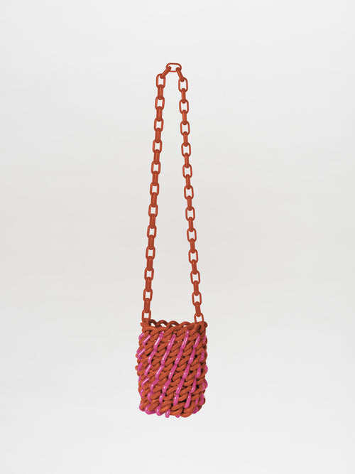 The Anise Bag Coral Magenta by SilviaTcherassi is a small, cylindrical accessory with a woven design in coral and magenta. It comes with a long chain-like strap, subtle resin rings, and is set against a plain white background.