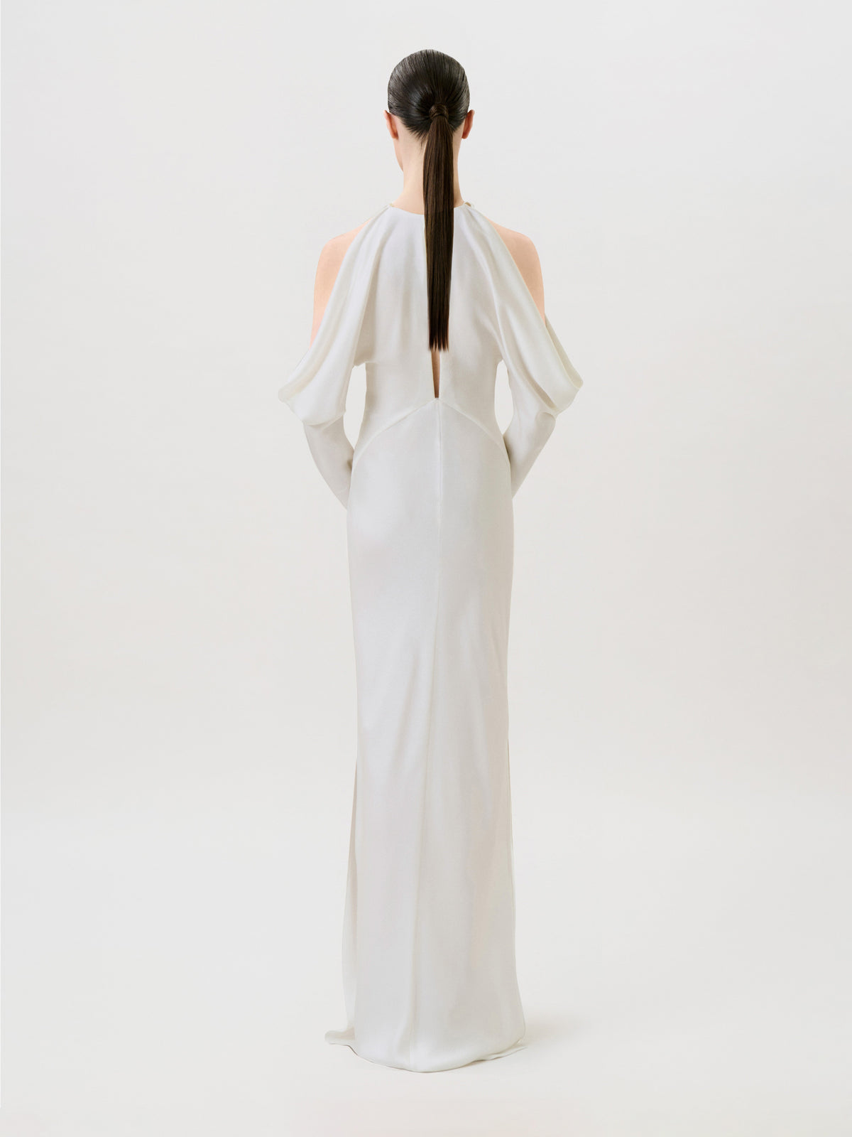 A woman with straight dark hair in a ponytail models the Antonia Dress, crafted in hammered satin and designed with open shoulders. Posed with her back to the camera against a simple light background, she captures the essence of modern ready-to-wear sophistication.