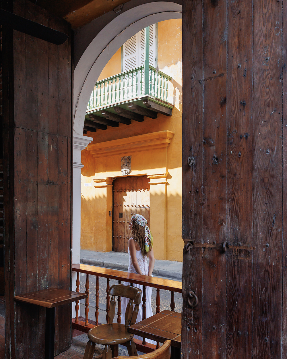 Woman standing in a sunlit courtyard with yellow walls and wooden doors.