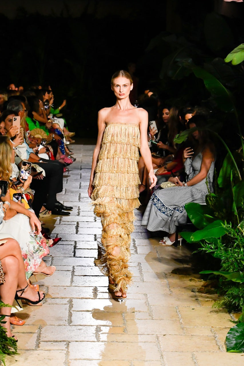 A model walks down a runway wearing a sleeveless, tiered, gold fringe dress. The setting is surrounded by greenery, with seated audience members on both sides. The floor is made of large stone tiles.