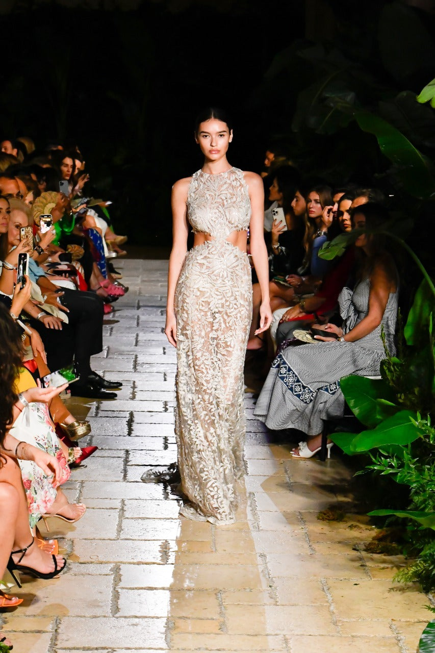 A model walks down a runway in a detailed, form-fitting, sleeveless gown with a cutout at the waist. The setting is lush, with greenery along the sides. The audience is seated close, capturing photos and observing the fashion show.