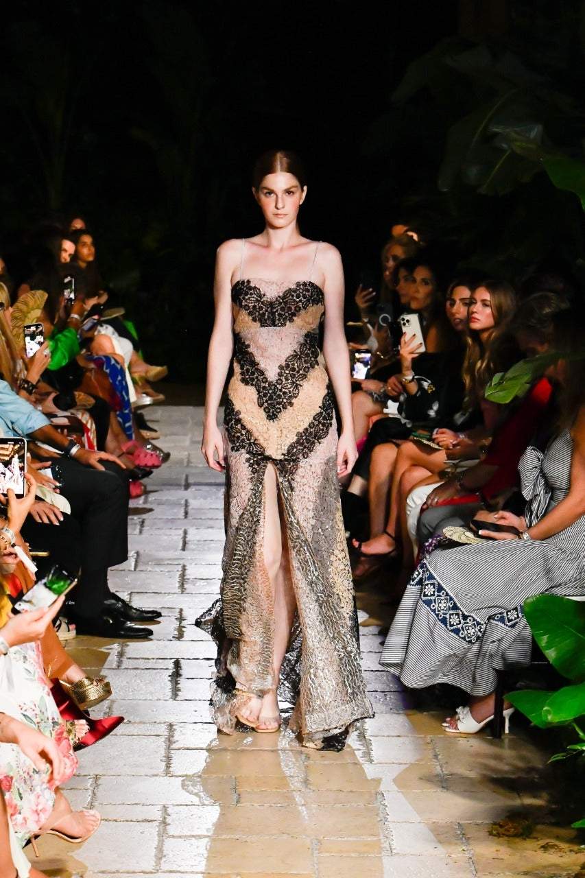 A model walks down a fashion runway wearing a sheer dress with intricate black designs. The audience is seated on either side, some holding phones to capture the moment. The setting appears to be a tropical or garden-themed environment.