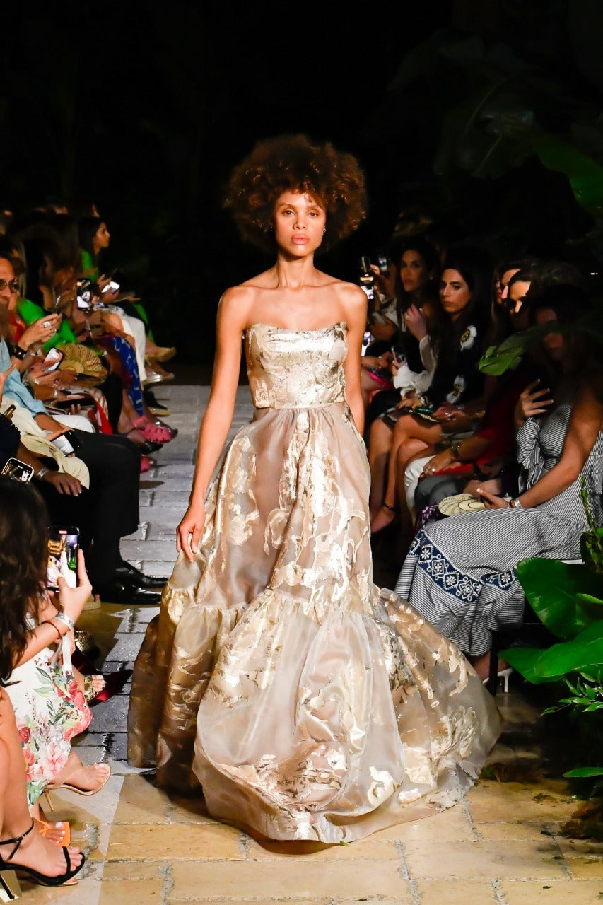 A model walks down a runway wearing a strapless, gold embroidered gown. The audience is seated along the sides, many holding phones to capture the event. The setting features tropical plants, suggesting an outdoor or garden location.