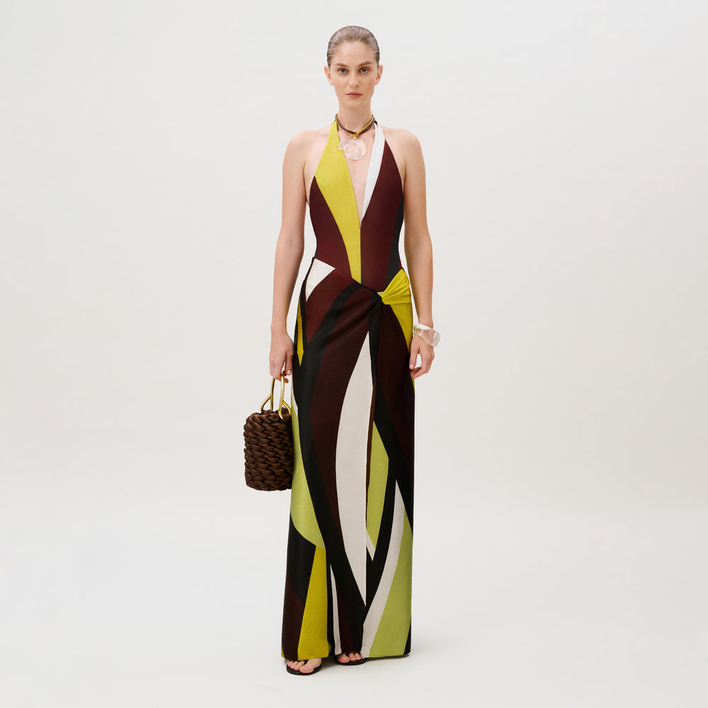 A woman wears an Aura Skirt floor-length halter dress with a geometric, multicolored abstract print made in Colombia and holds a dark brown woven handbag.