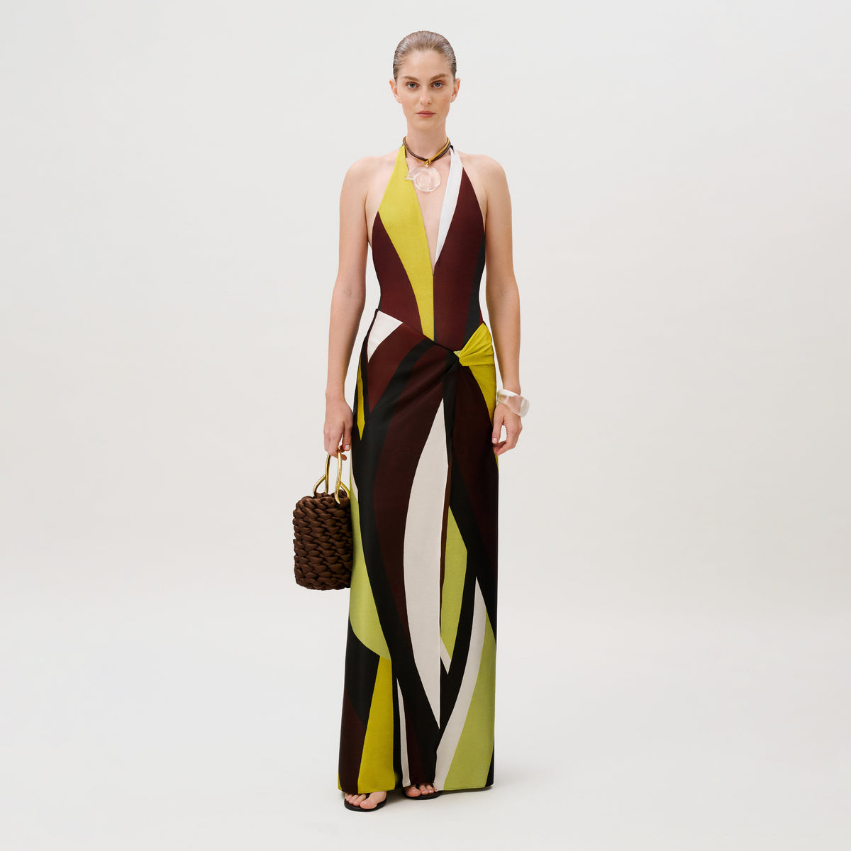 A woman wears an Aura Skirt floor-length halter dress with a geometric, multicolored abstract print made in Colombia and holds a dark brown woven handbag.