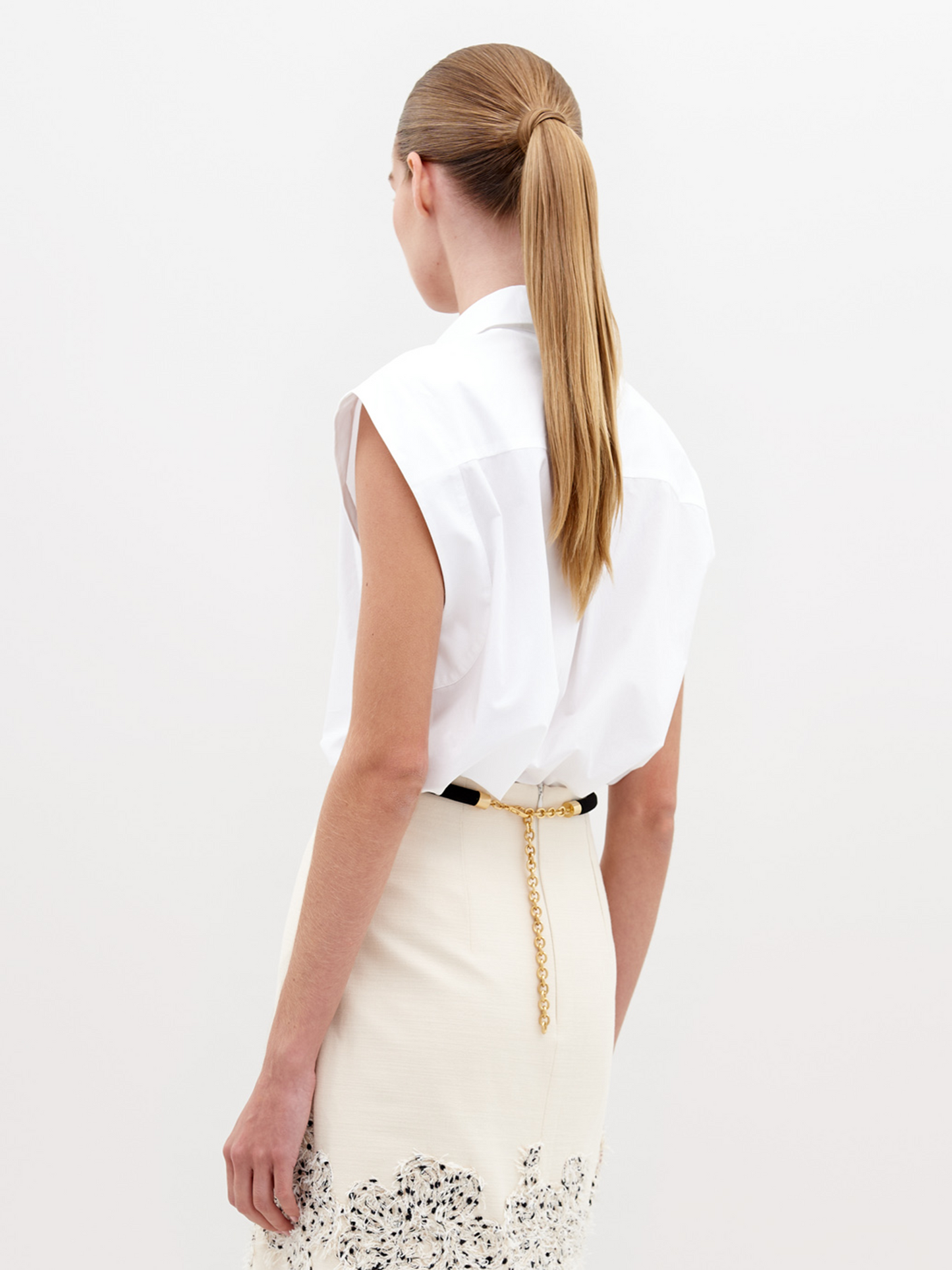 A woman wearing the Azalea Blouse White with floral decorations and a light beige skirt with a black belt and lace detailing stands against a plain background. She holds a small dark bag, showcasing the chic ensemble set to ship by date, November 15th, 2024.