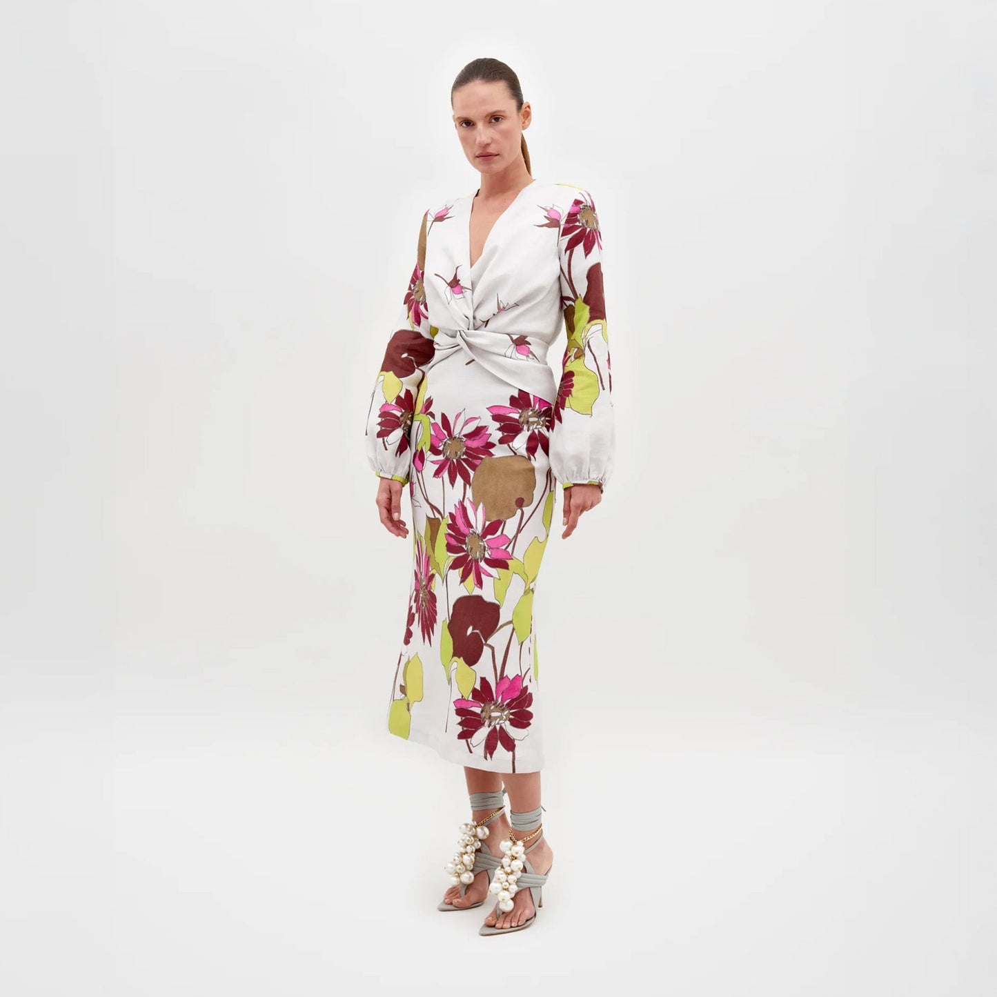 A woman stands against a plain background wearing the Battia Dress in Botanical Multifloral, which features a bold floral print, a cinched waist, and long sleeves. She is also wearing gray high-heeled sandals adorned with large pearl-like embellishments. The dress is available for pre-order now, with an estimated shipping date of February 15th, 2025.