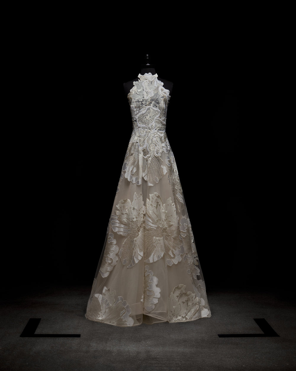 The Becca Dress by SVT DESIGNS, displayed on a mannequin against a dark background, features intricate white floral lace patterns over sheer fabric, creating an elegant and delicate appearance.